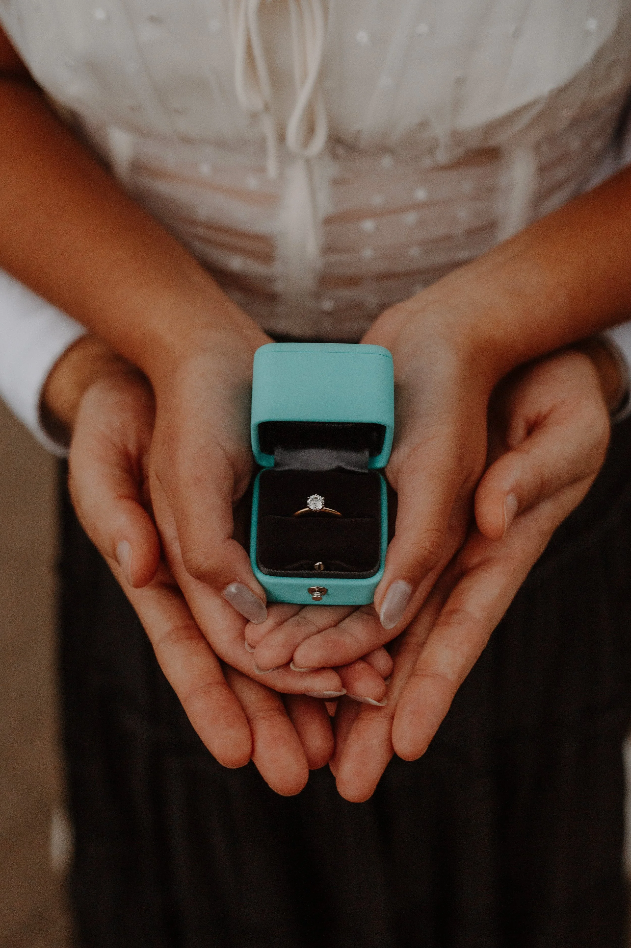 Close-up of hands holding an open teal ring box with a diamond engagement ring