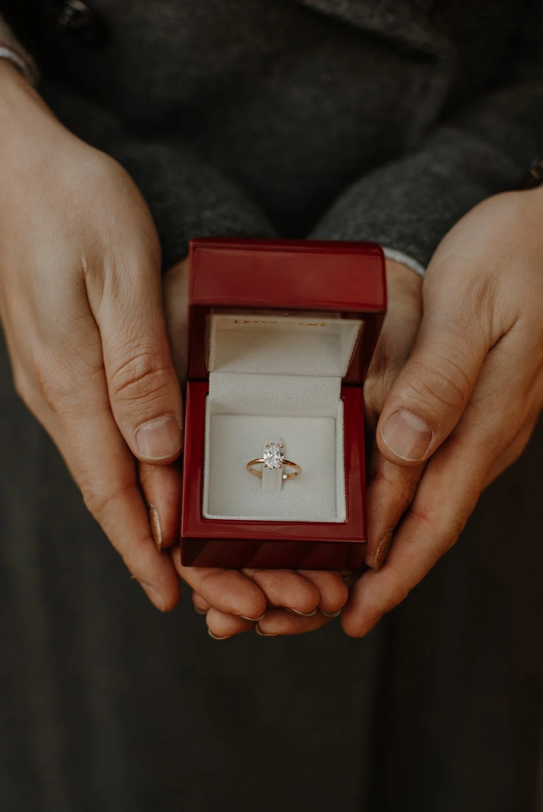 Close-up of a solitaire engagement ring in a red box held by a newly engaged couple