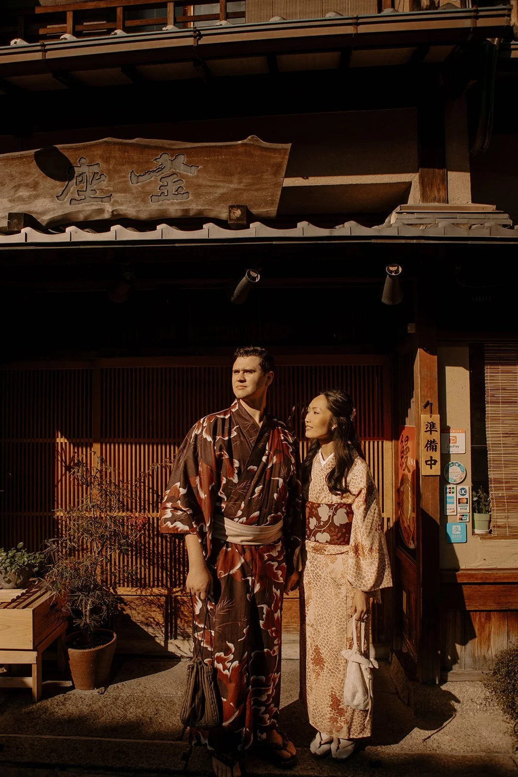 Couple in kimono standing in golden evening light outside a traditional wooden storefront in Kyoto