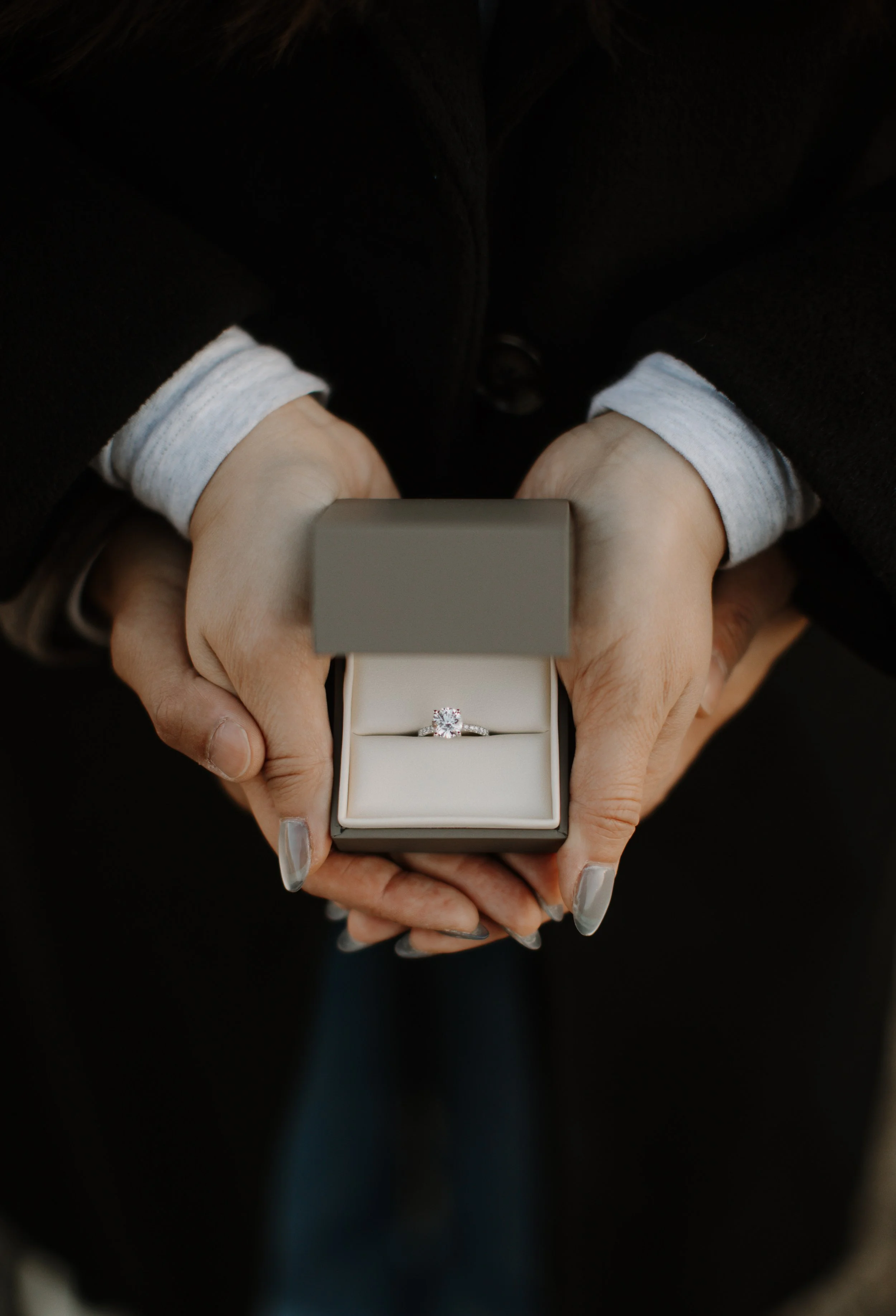 Close-up of hands holding an open ring box with a diamond engagement ring against a dark background