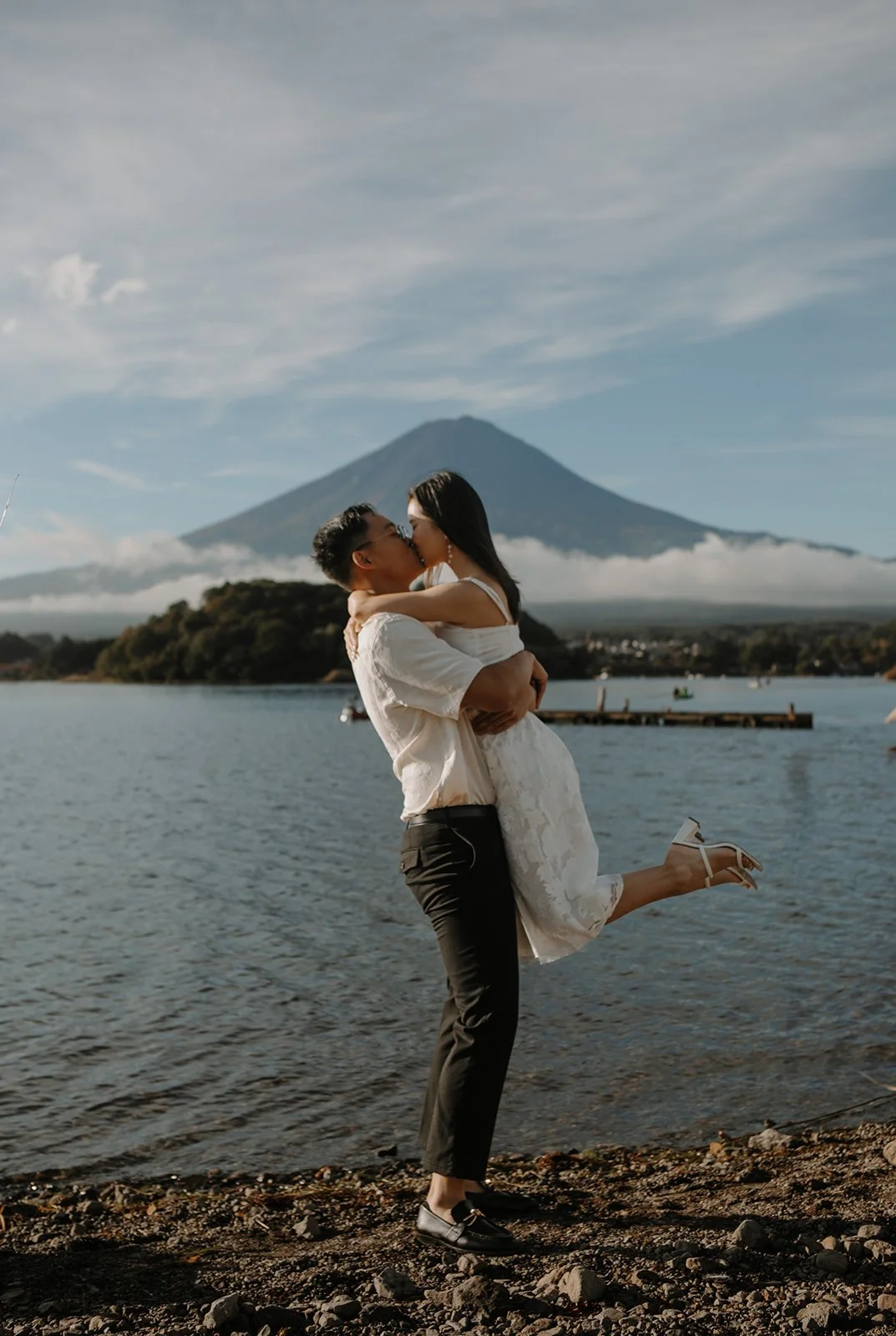 Couple shares a playful lifted kiss beside a wooden dock on Lake Kawaguchiko, Mount Fuji behind them.