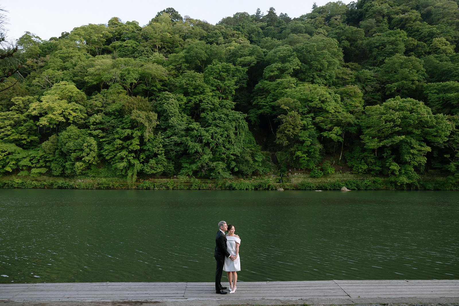 Couple embraces on a wooden dock beside a wide green river in the Kyoto hills.