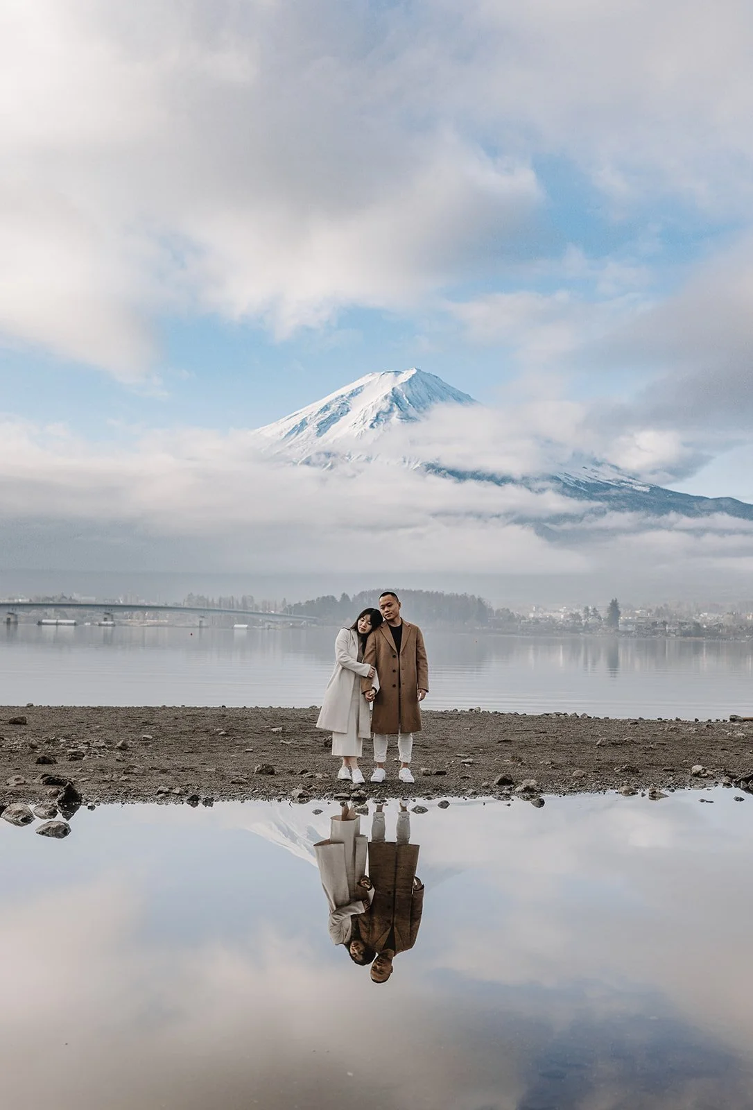 Couple stands close together on the shoreline, Mount Fuji and their reflection visible in the still water.