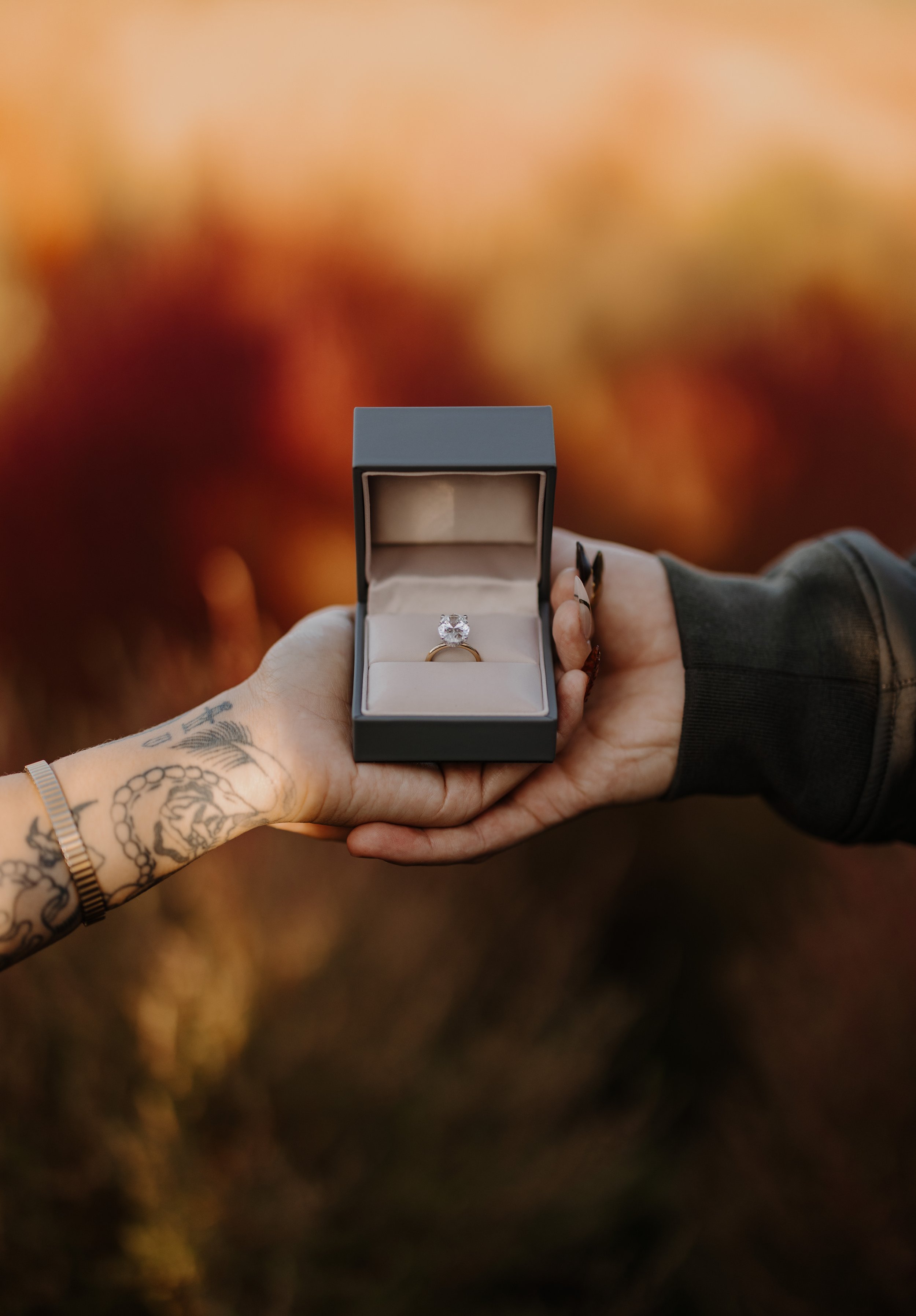 Close-up of a tattooed hand presenting an open ring box with a diamond engagement ring in an autumn field