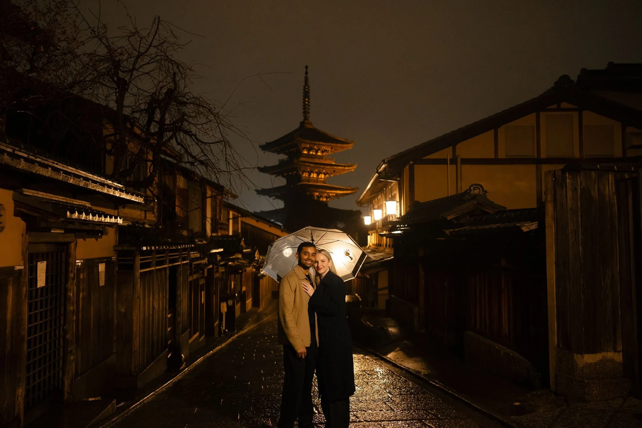 Couple sharing an umbrella on a rainy cobblestone street in Gion with Yasaka Pagoda lit up behind them