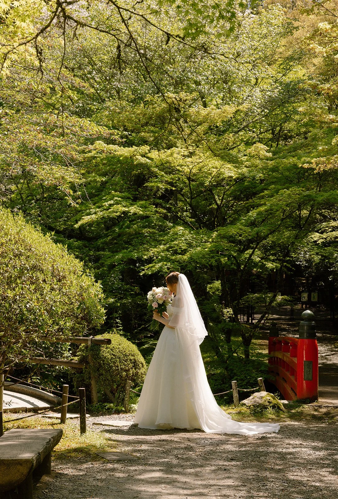 Bride in a white gown and veil pauses beside a small red bridge in a Kyoto garden.