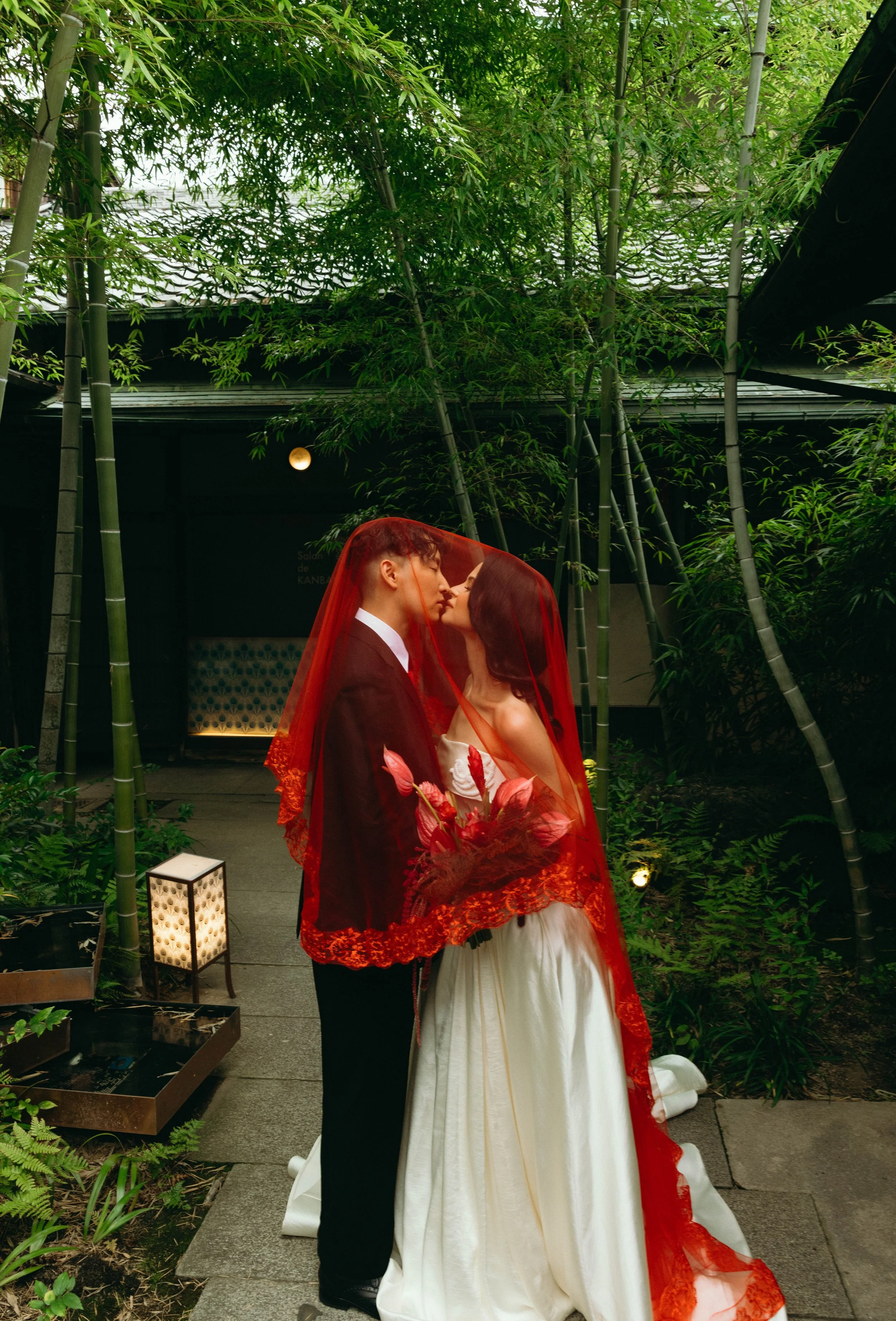 Bride and groom share a kiss beneath a red veil in a lantern-lit bamboo garden at night.