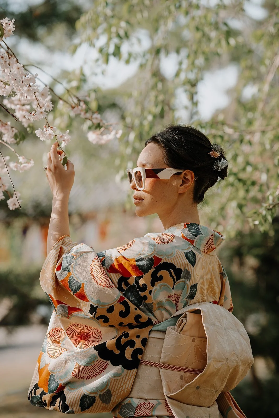 Woman in a floral kimono reaching up to cherry blossom branches in a Kyoto garden