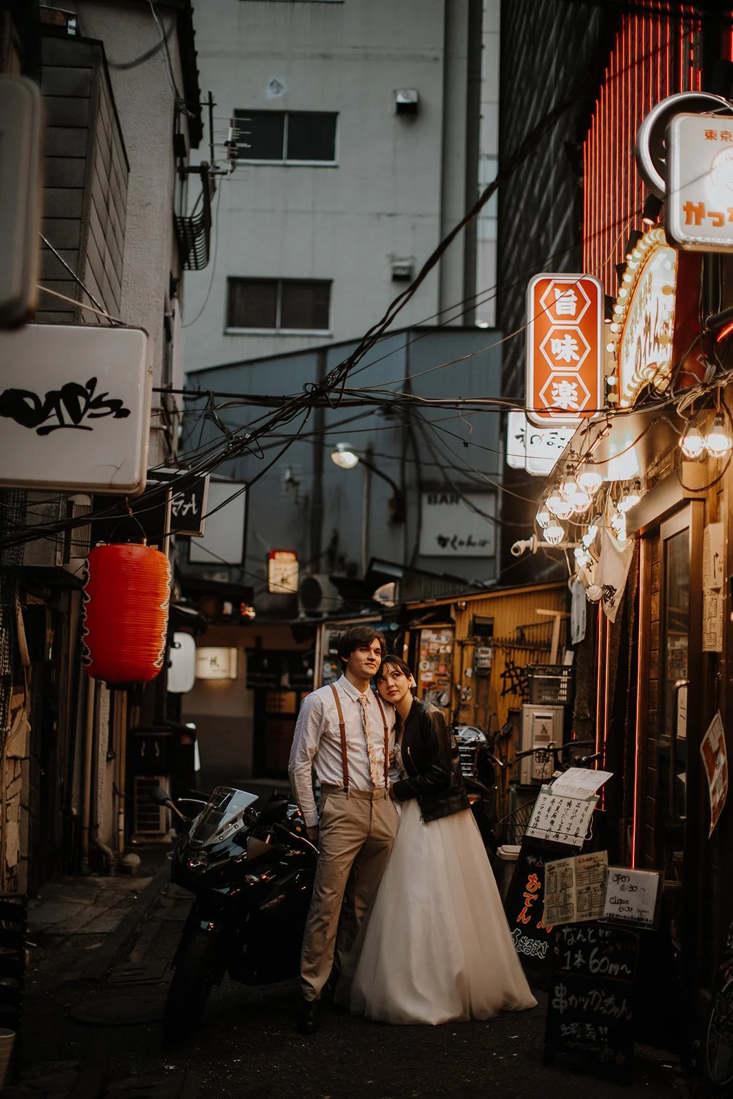 Couple embraces beside a motorcycle in a narrow, lantern-lit Tokyo alleyway at night.