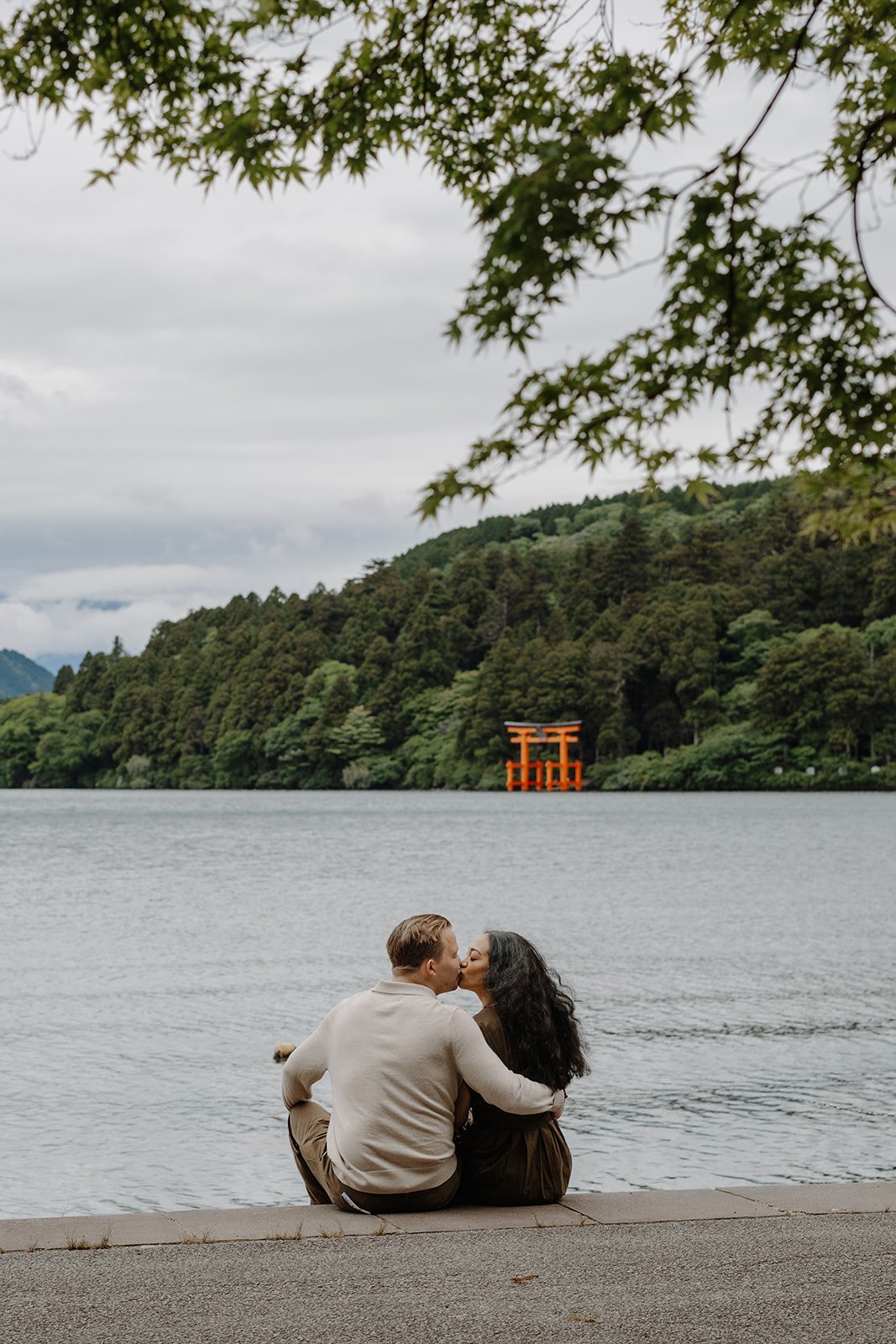 Couple sitting on the shore of Lake Ashi kissing, framed by maple branches, with the red torii gate visible across the water, Hakone