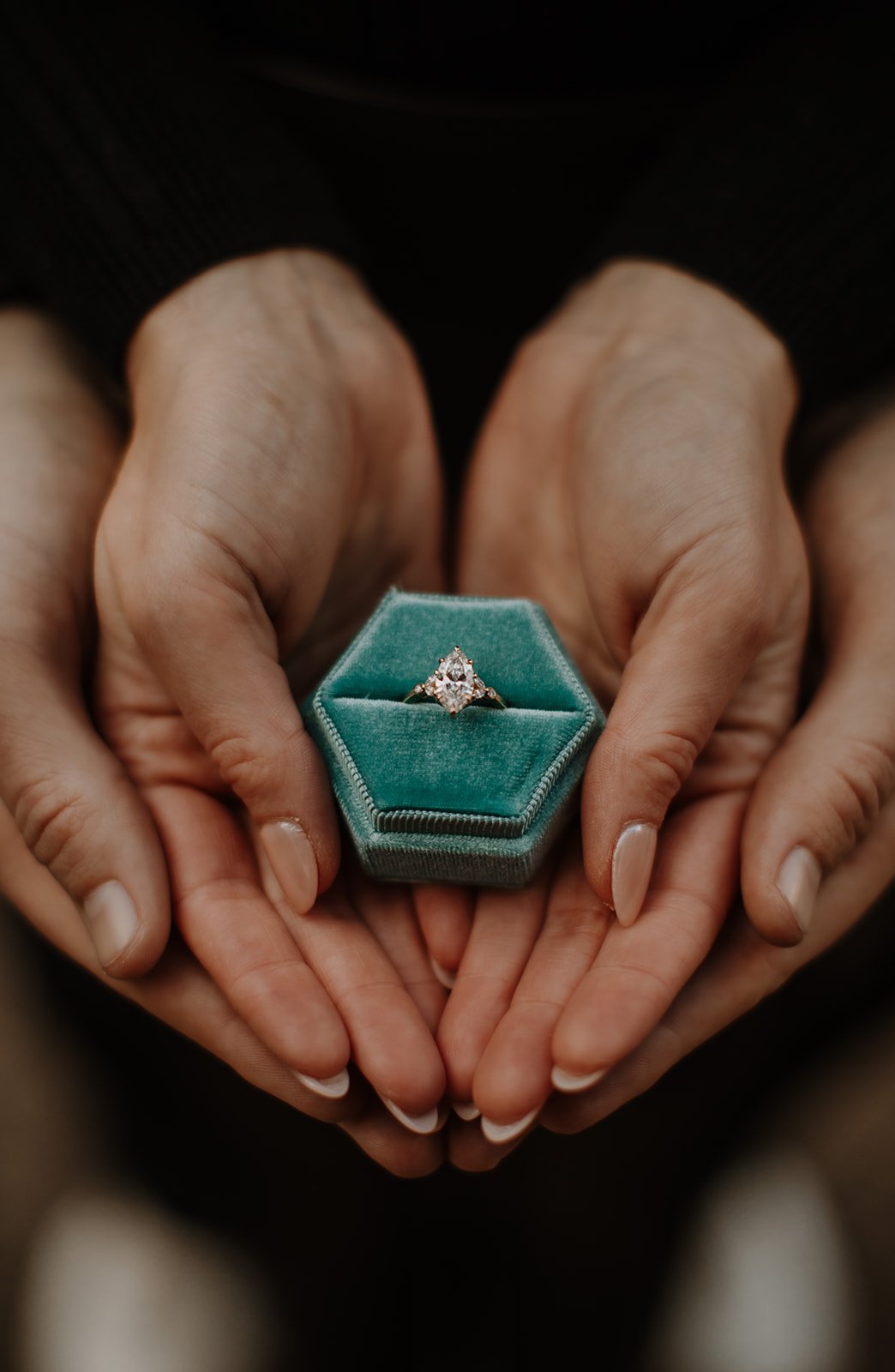 Detail shot of an engagement ring in a teal velvet box, framed by the couple's hands forming a heart.