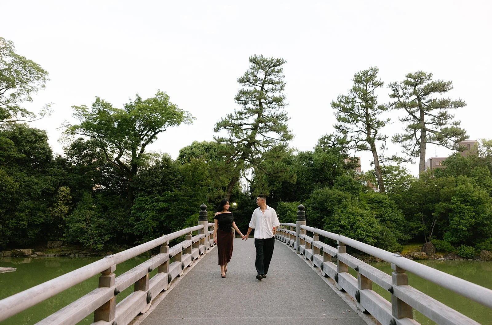 Newly engaged couple walking hand in hand toward the camera across a wooden bridge in Kyoto
