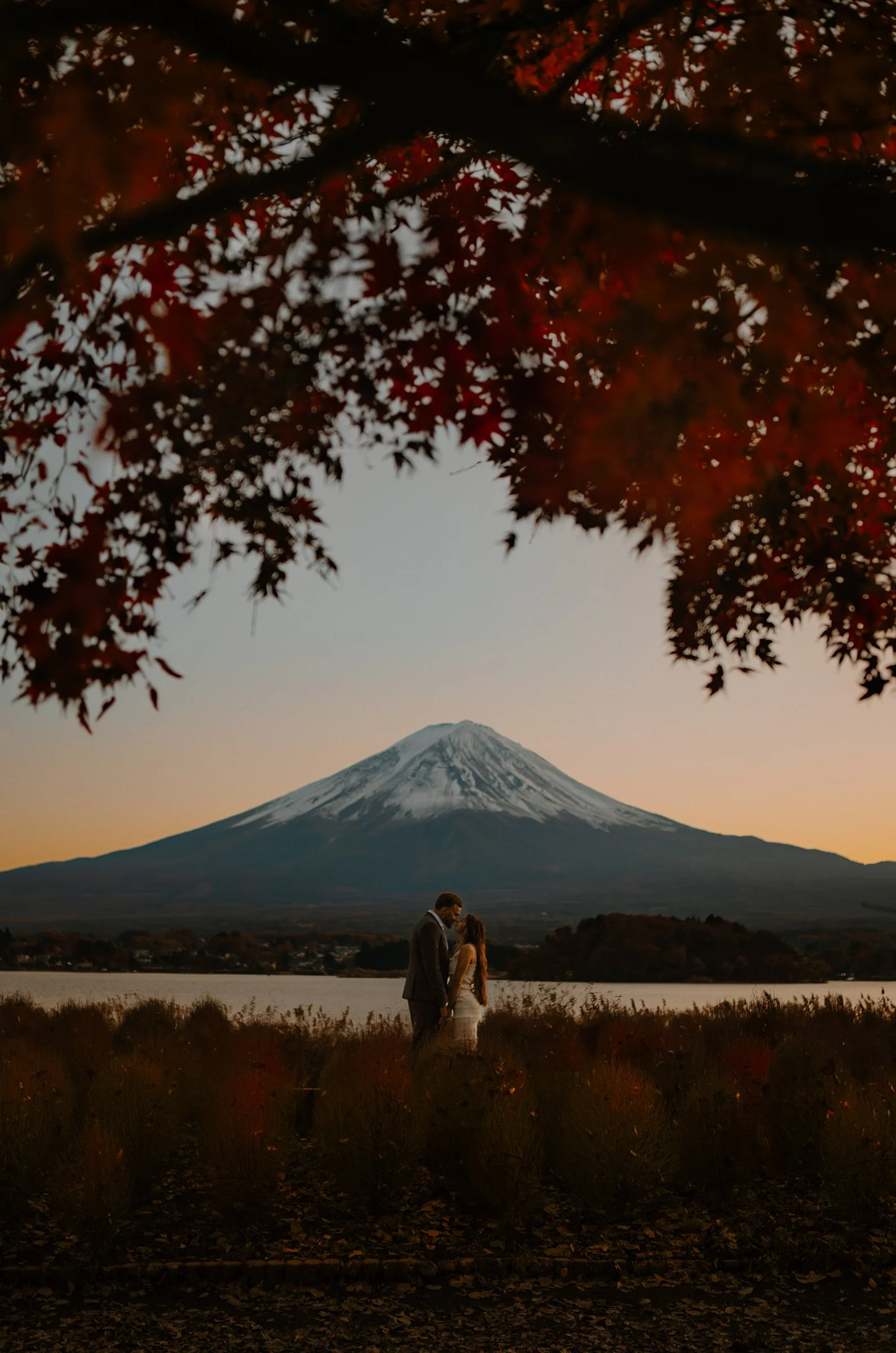 Bride and groom embracing before snow-capped Mount Fuji and Lake Kawaguchi, framed by autumn maple leaves at sunset, during a Japan elopement