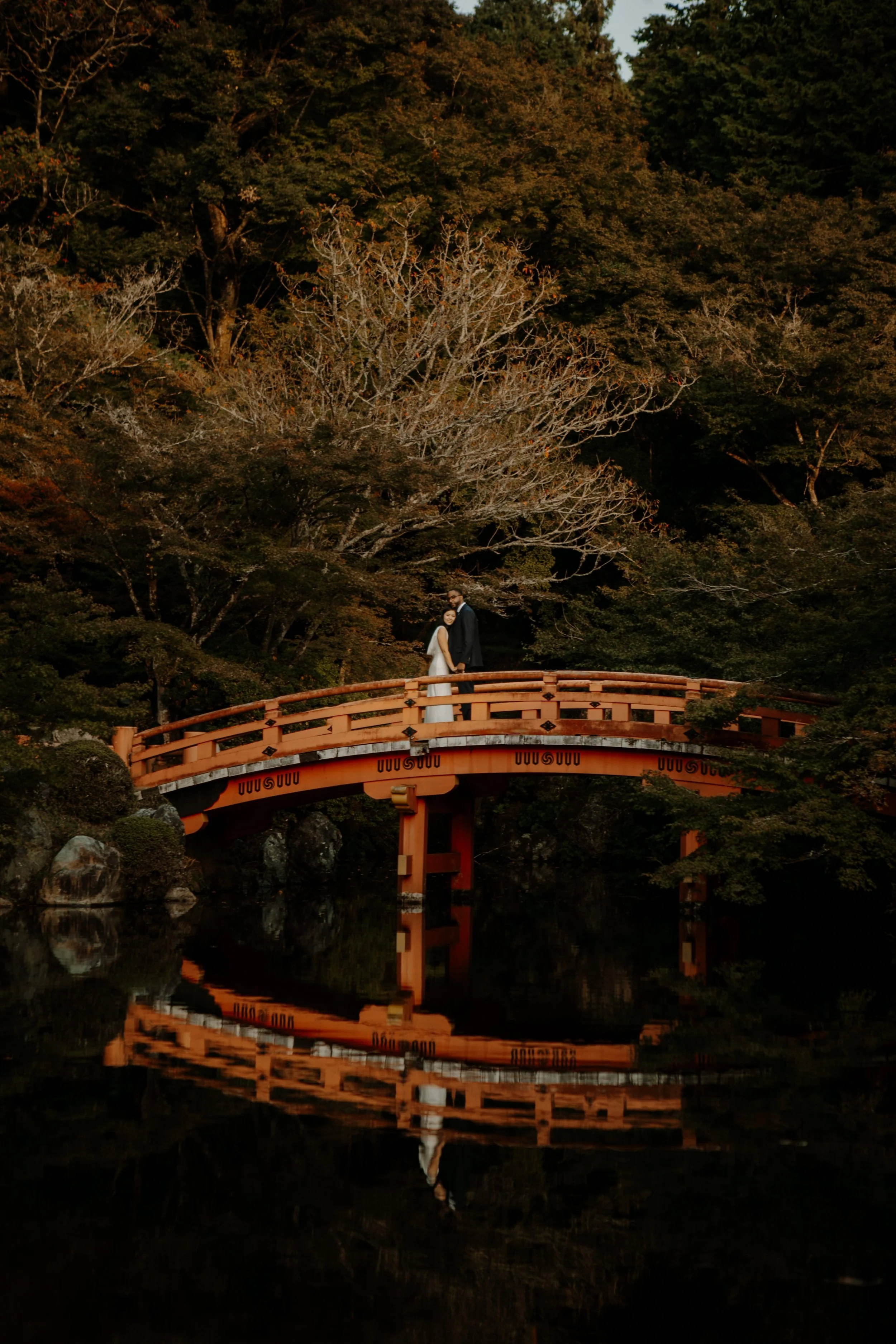 Bride and groom standing on Daigoji Temple's red bridge in Kyoto, their reflection mirrored in the still pond below