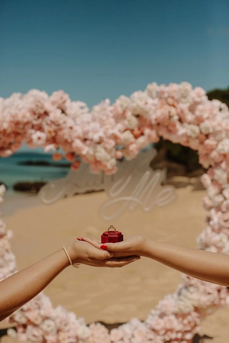 Close-up of hands exchanging a red ring box beneath a flower arch reading "Marry Me" on an Okinawa beach