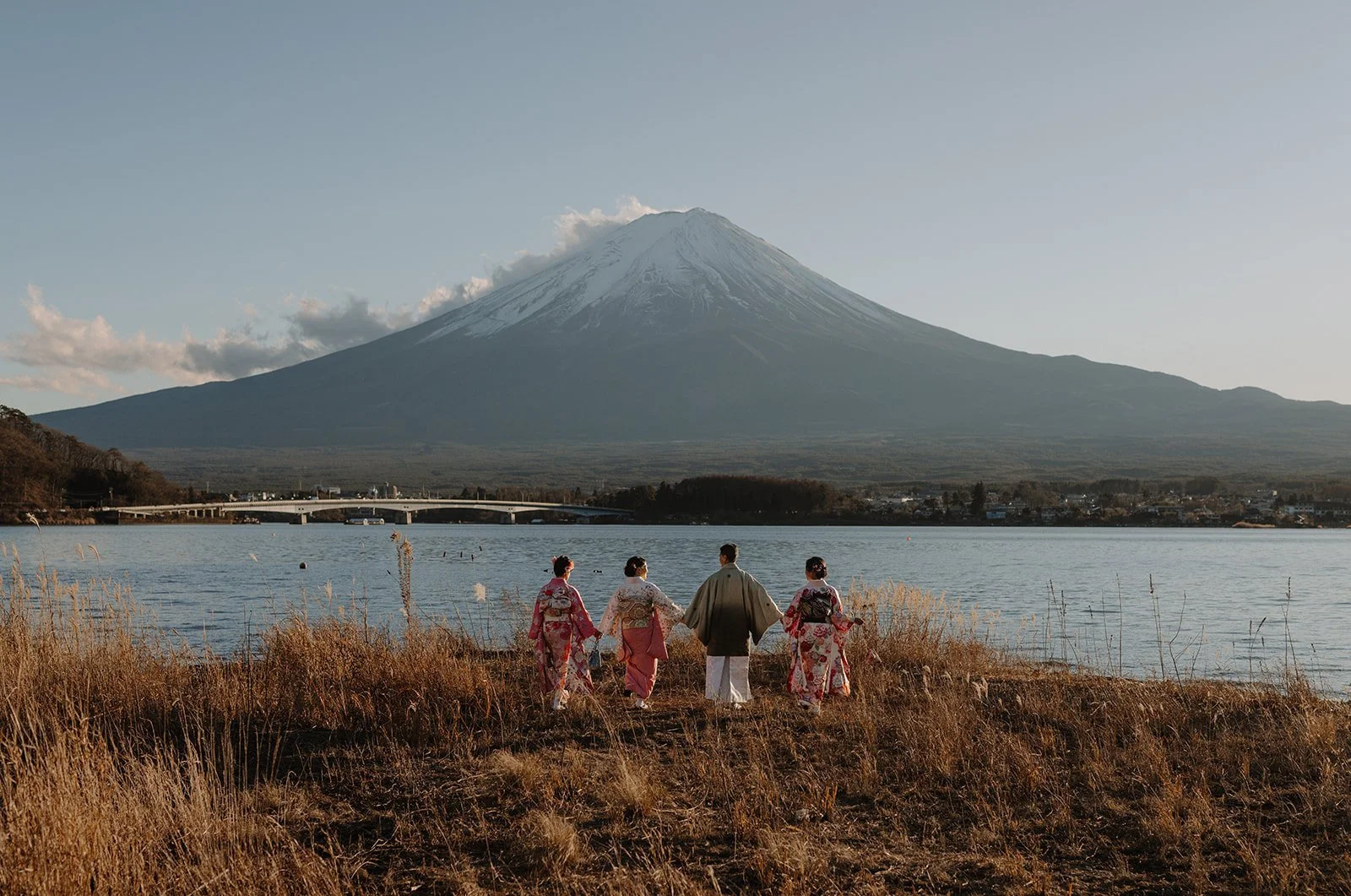 Family in kimono holding hands and walking along the grassy shoreline of Lake Kawaguchiko with Mount Fuji in the background