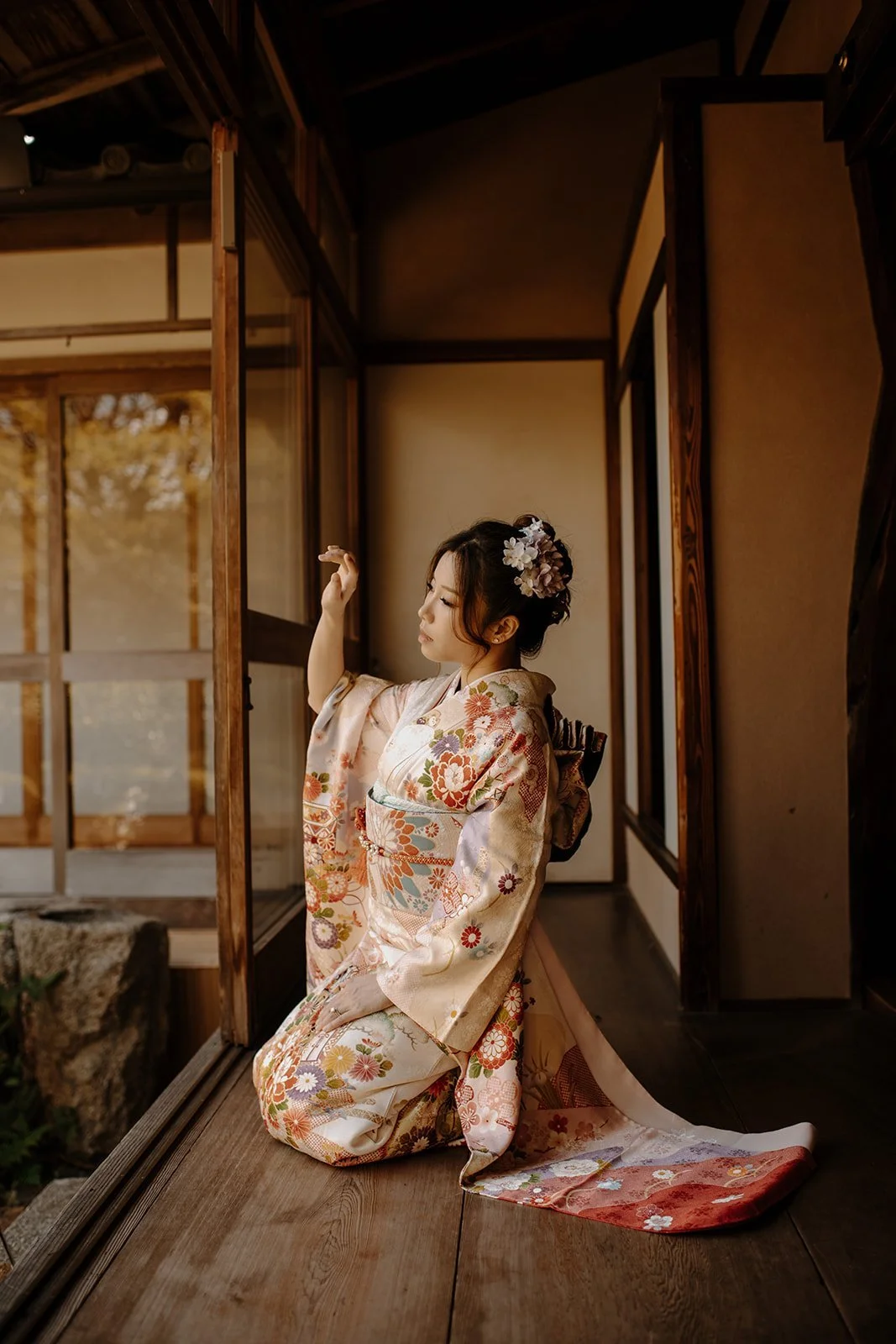 Woman in an ivory floral kimono kneeling on a wooden engawa porch, reaching up toward the light, in a traditional Kyoto house