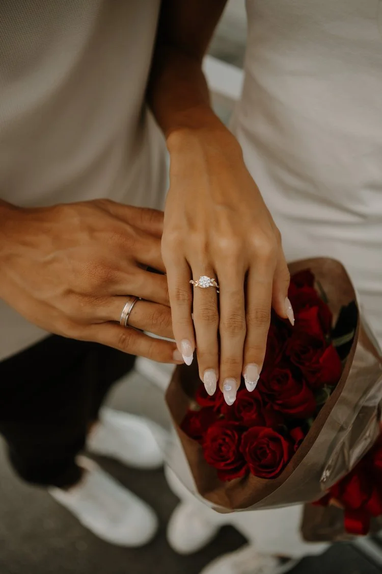 Close-up of a couple's clasped hands with an engagement ring, resting on a bouquet of red roses