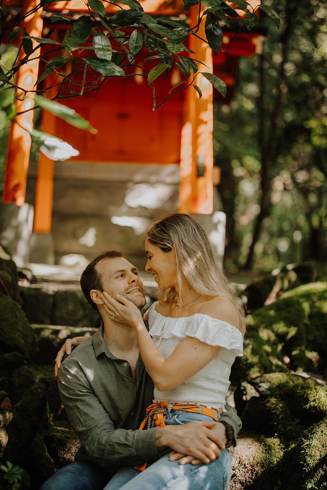 Couple sitting on moss-covered stone steps beneath a small red torii gate in the forest approach to Hakone Shrine