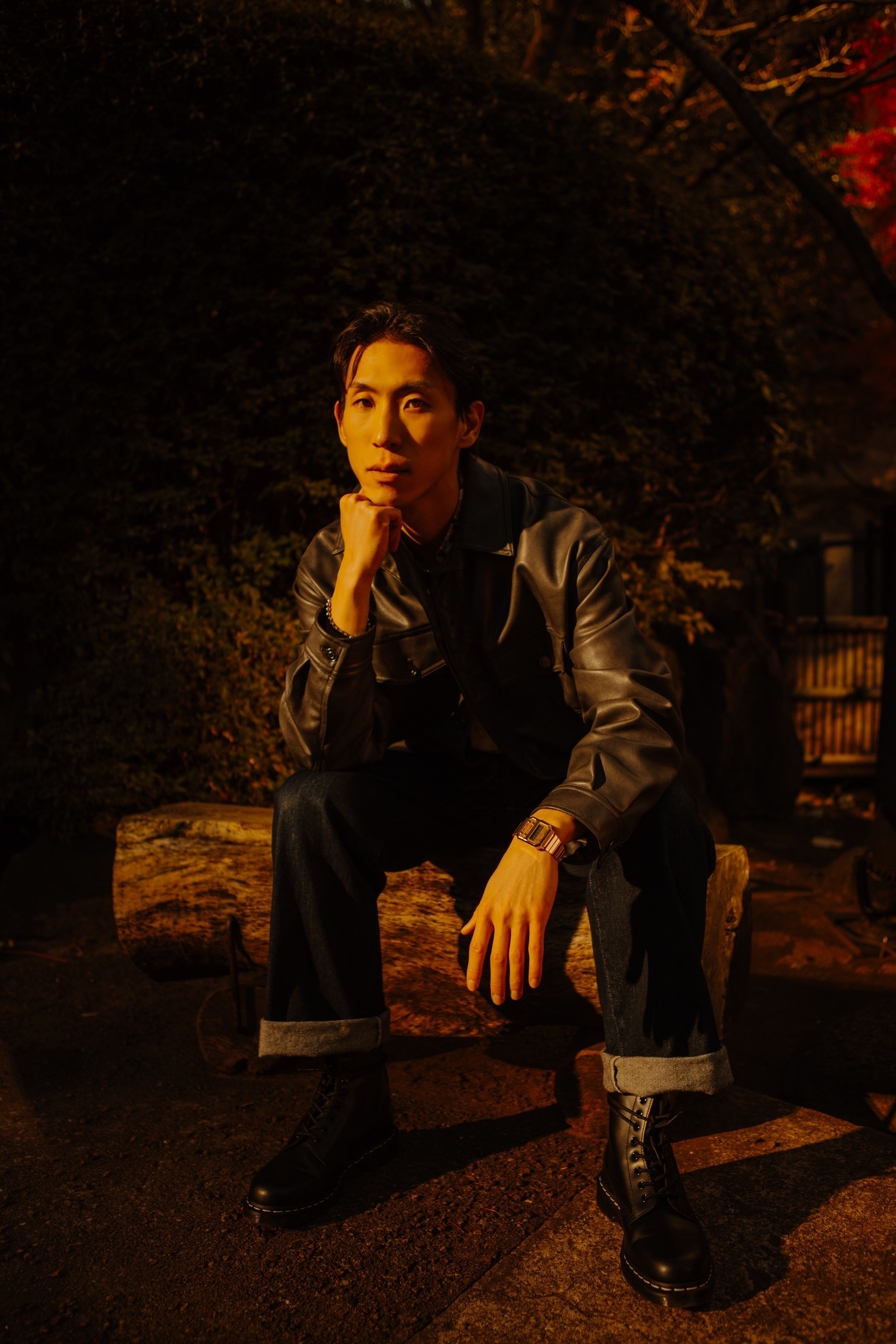 Man in a leather jacket seated on a log in a Tokyo garden surrounded by autumn maple leaves.