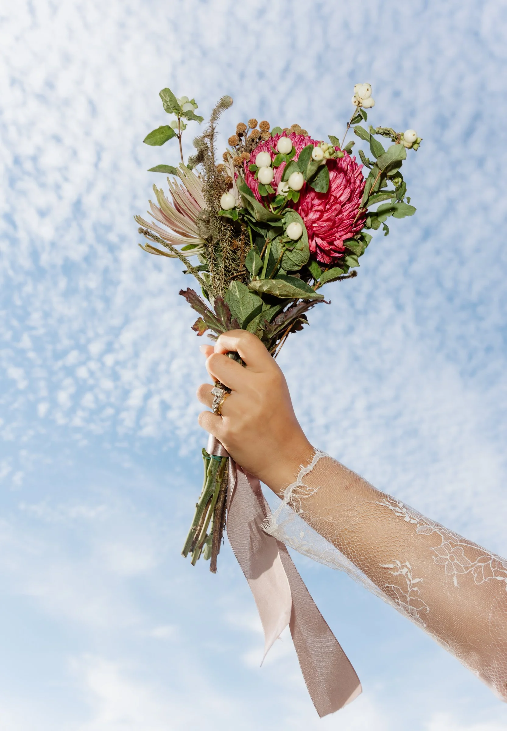 Bride's lace-sleeved arm raising a bouquet against a blue sky with soft clouds in Japan