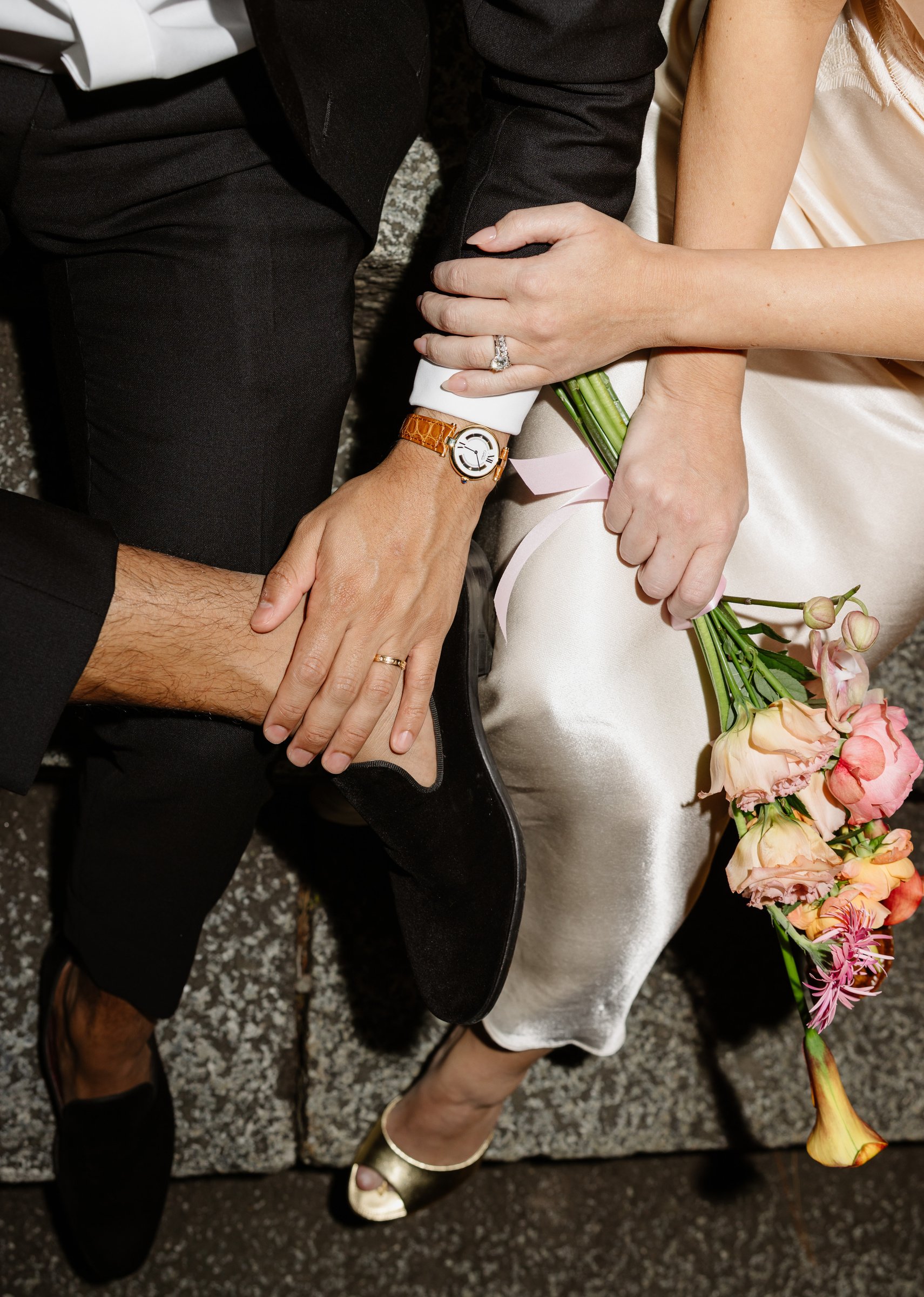 Close detail of a couple's joined hands with a wristwatch and a pink bouquet on stone steps in Japan