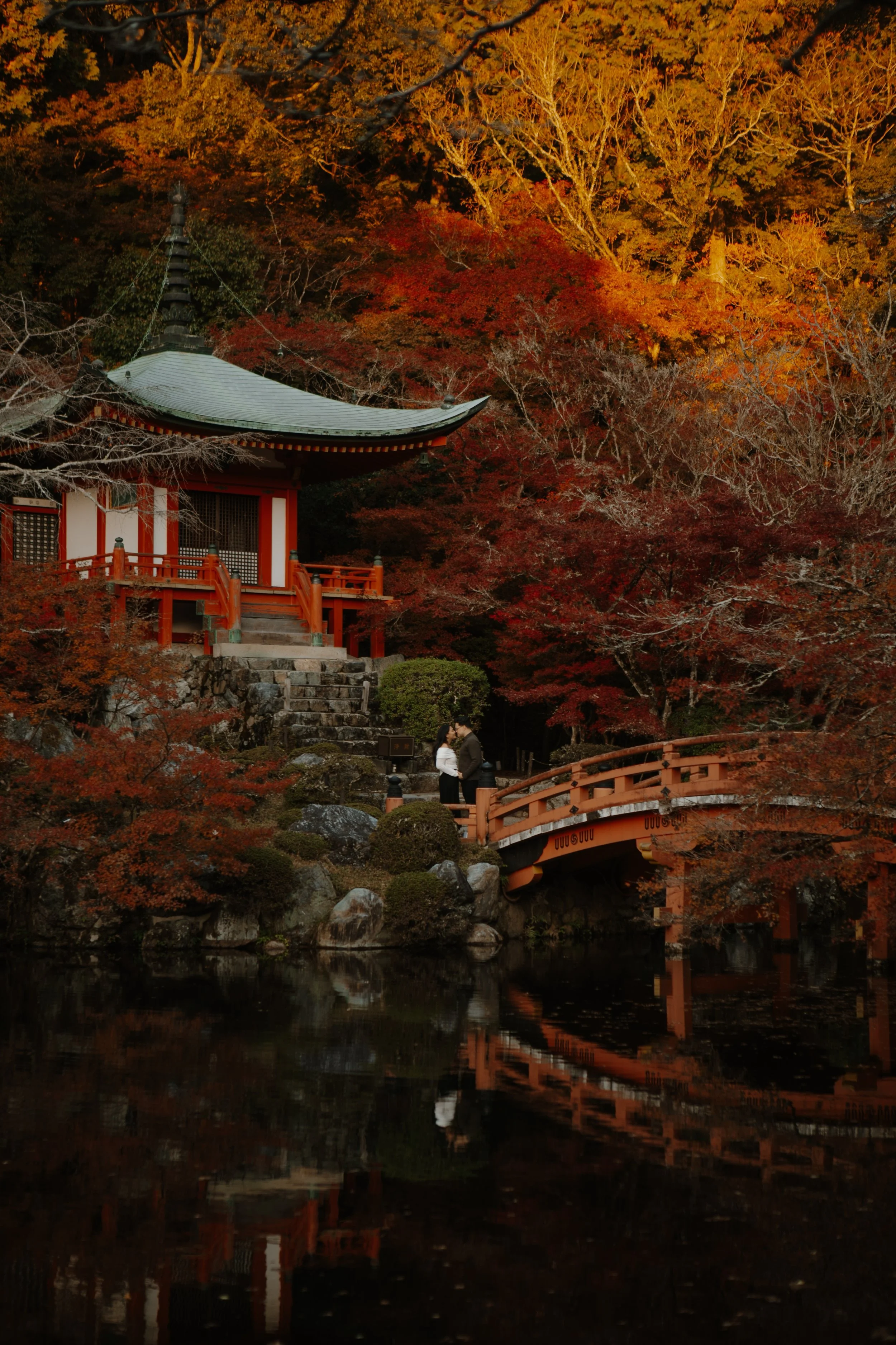 Wide autumn view of Daigoji Temple's Bentendo Hall and red bridge surrounded by fiery maple foliage, with a couple embracing below.