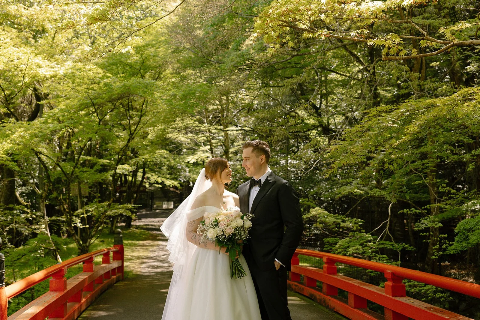 Couple standing close together on a red lacquered bridge under spring maples in Kyoto