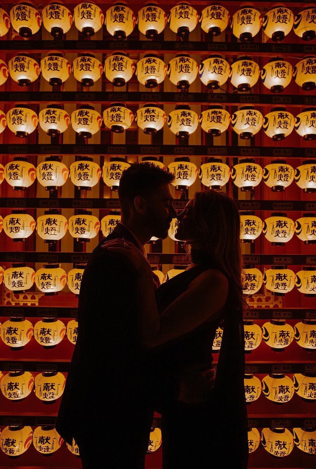 Silhouetted couple embracing in front of a glowing wall of paper lanterns at a Japanese shrine.