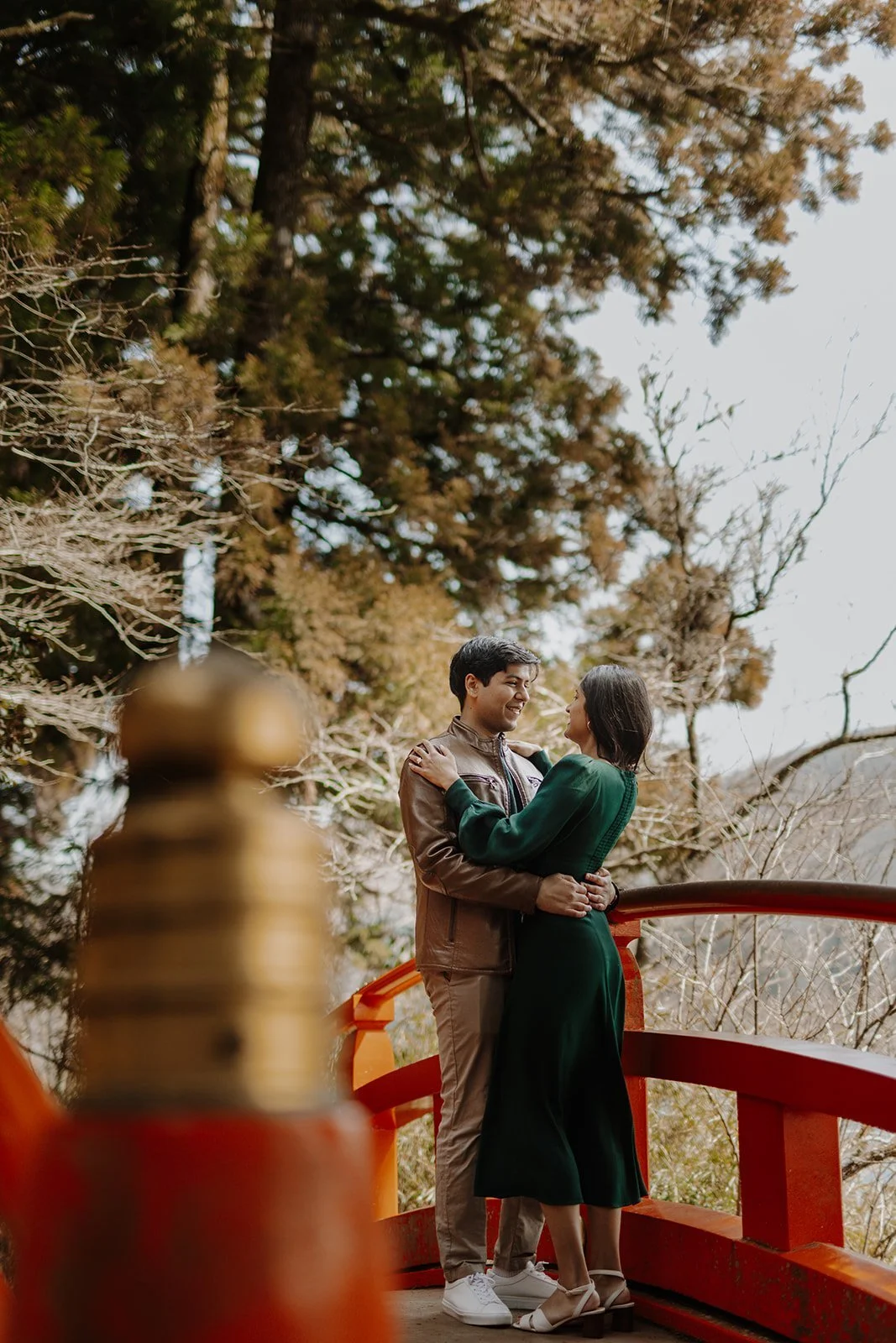 Newly engaged couple embracing on a red shrine bridge surrounded by trees, Hakone
