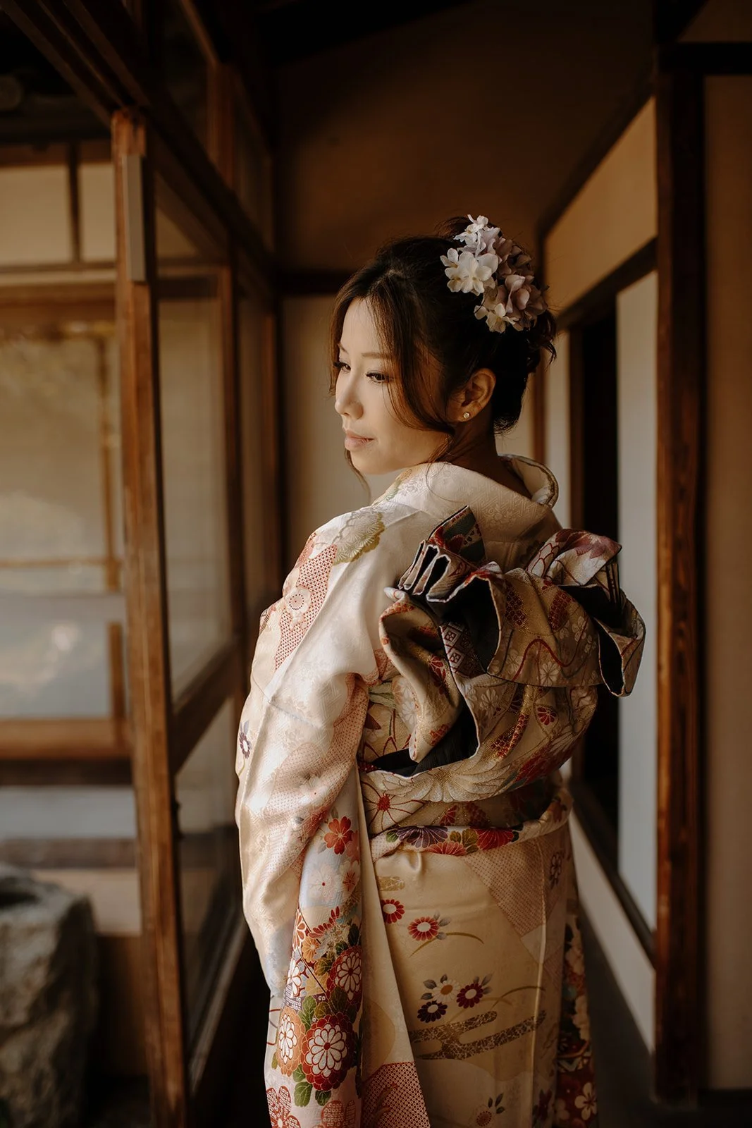 Rear view of a woman in an ivory floral kimono showing the elaborate obi bow, standing in a wooden temple corridor in Kyoto