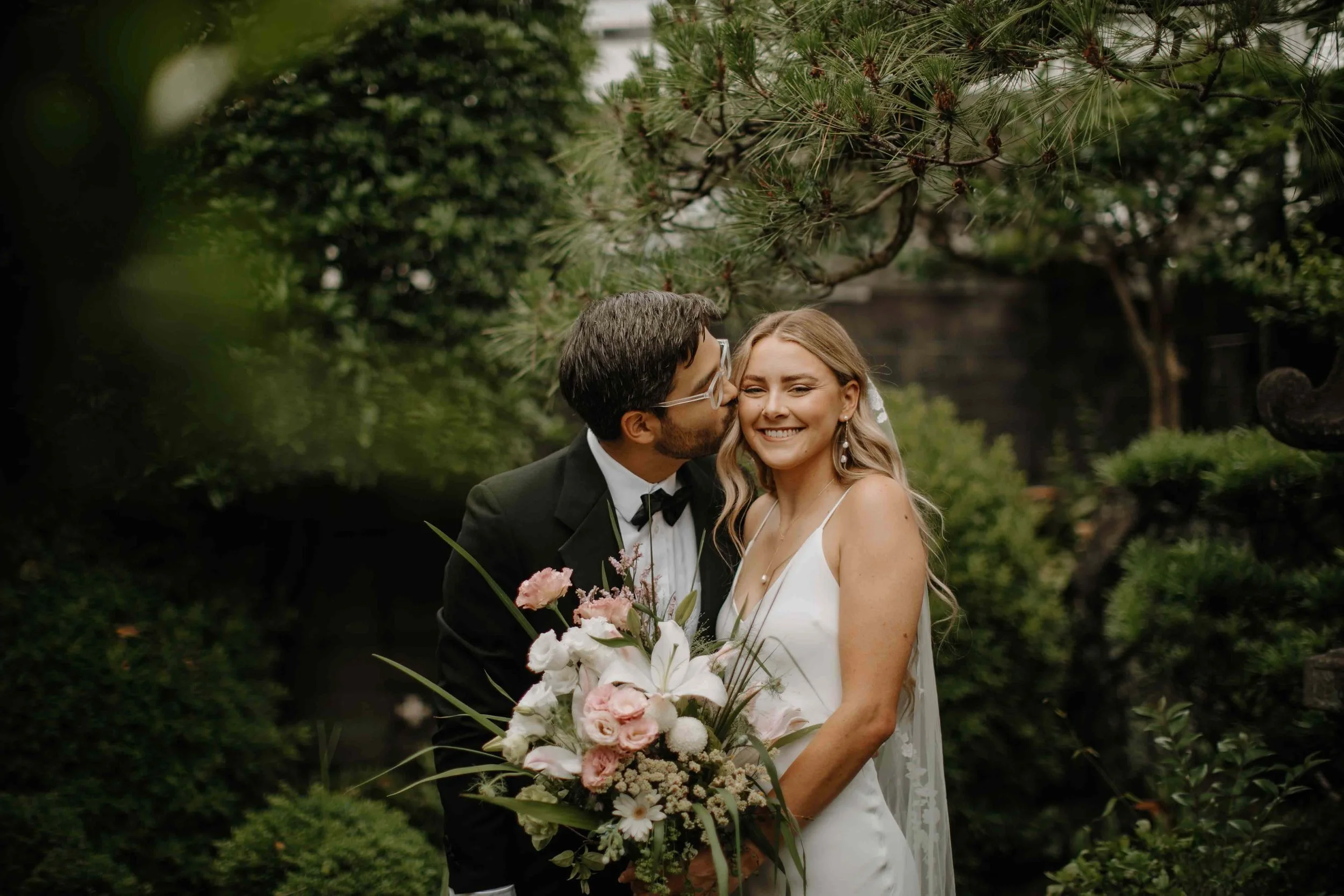 Groom kissing his bride's forehead in a green garden as she holds a pink and white bridal bouquet.