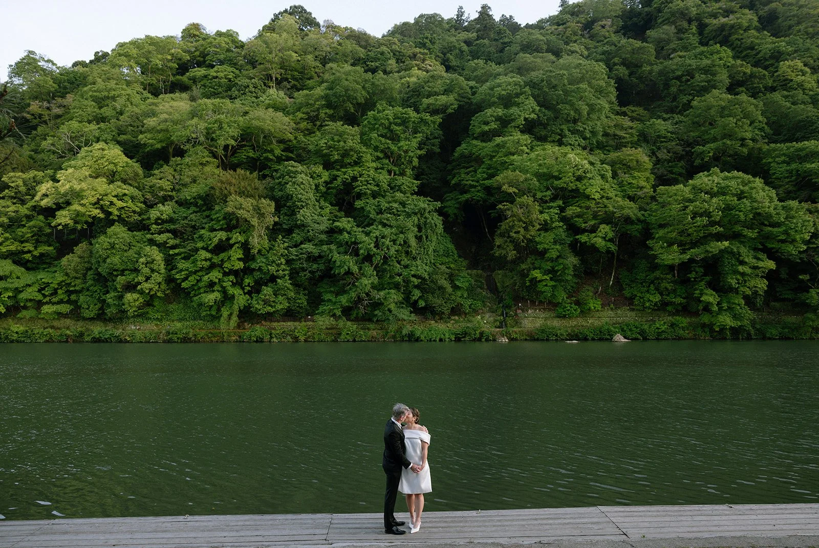 Bride and groom kiss on the riverside dock in Arashiyama, the forested hillside rising behind them.