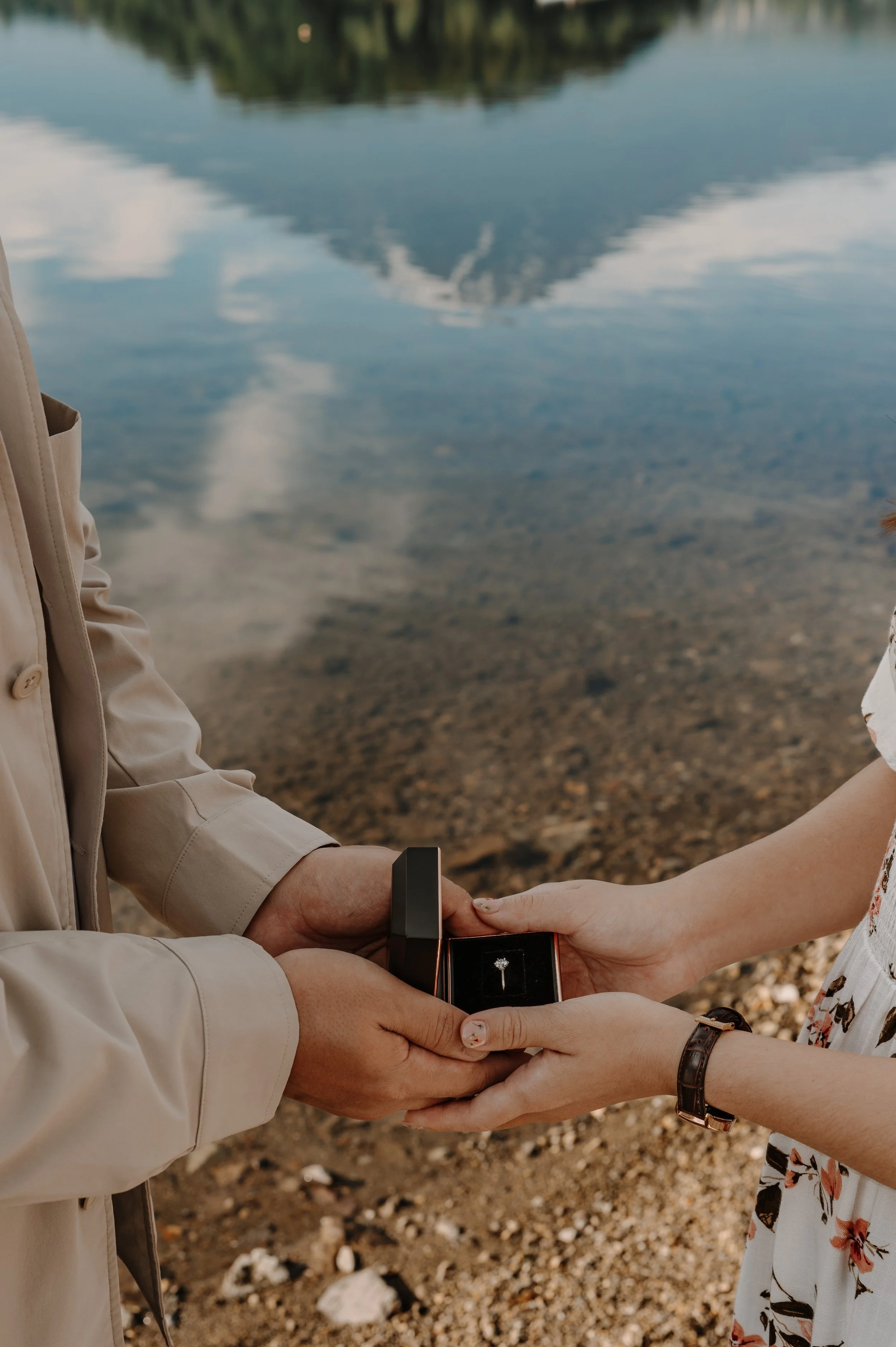 Close-up of hands holding an open ring box with a diamond ring beside a clear lakeshore