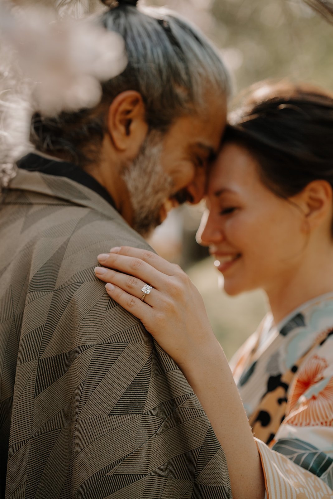 Couple in kimono touching foreheads beneath cherry blossoms, the bride's engagement ring visible on her hand.