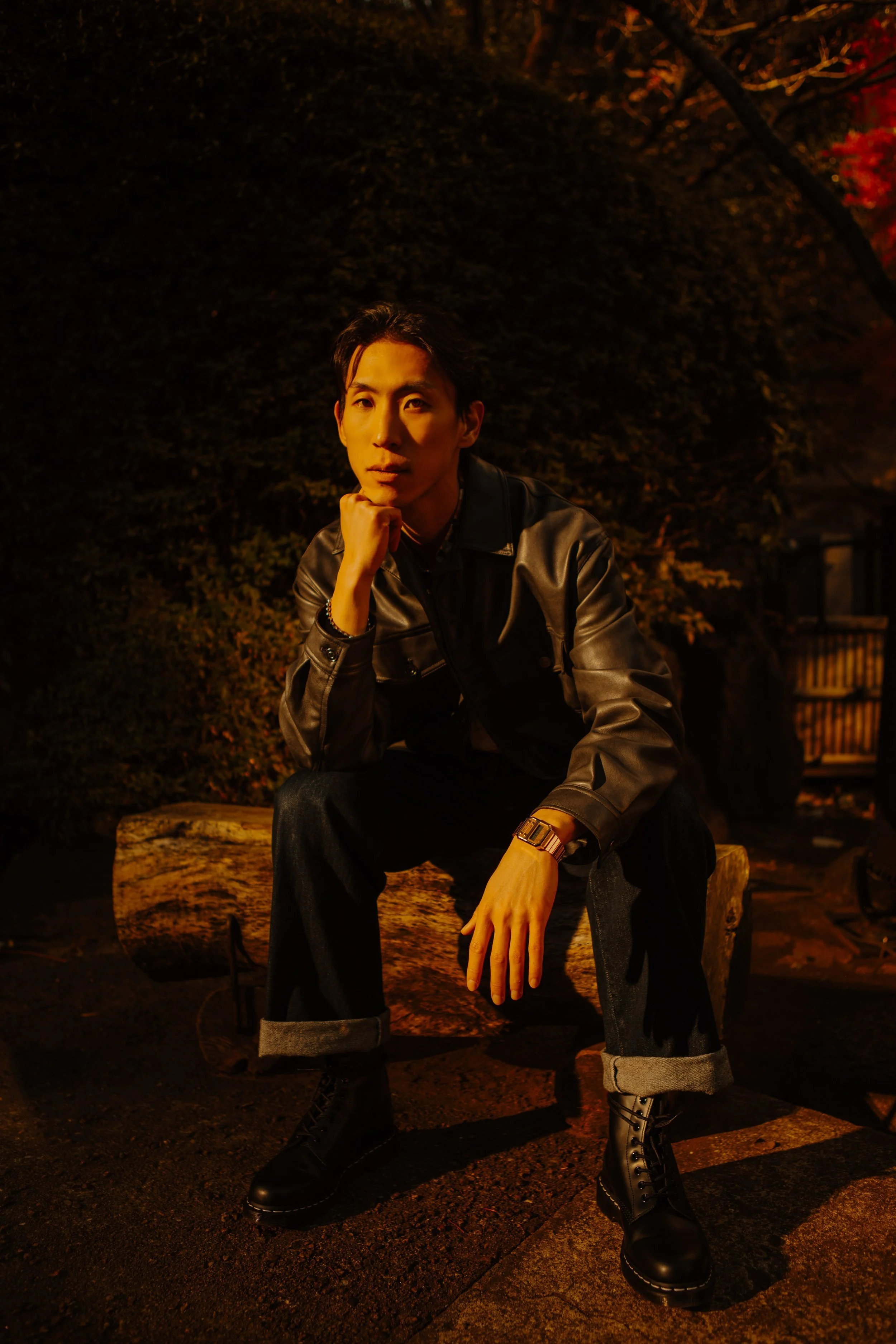 Man in a leather jacket seated on a log in a Tokyo garden surrounded by autumn maple leaves.