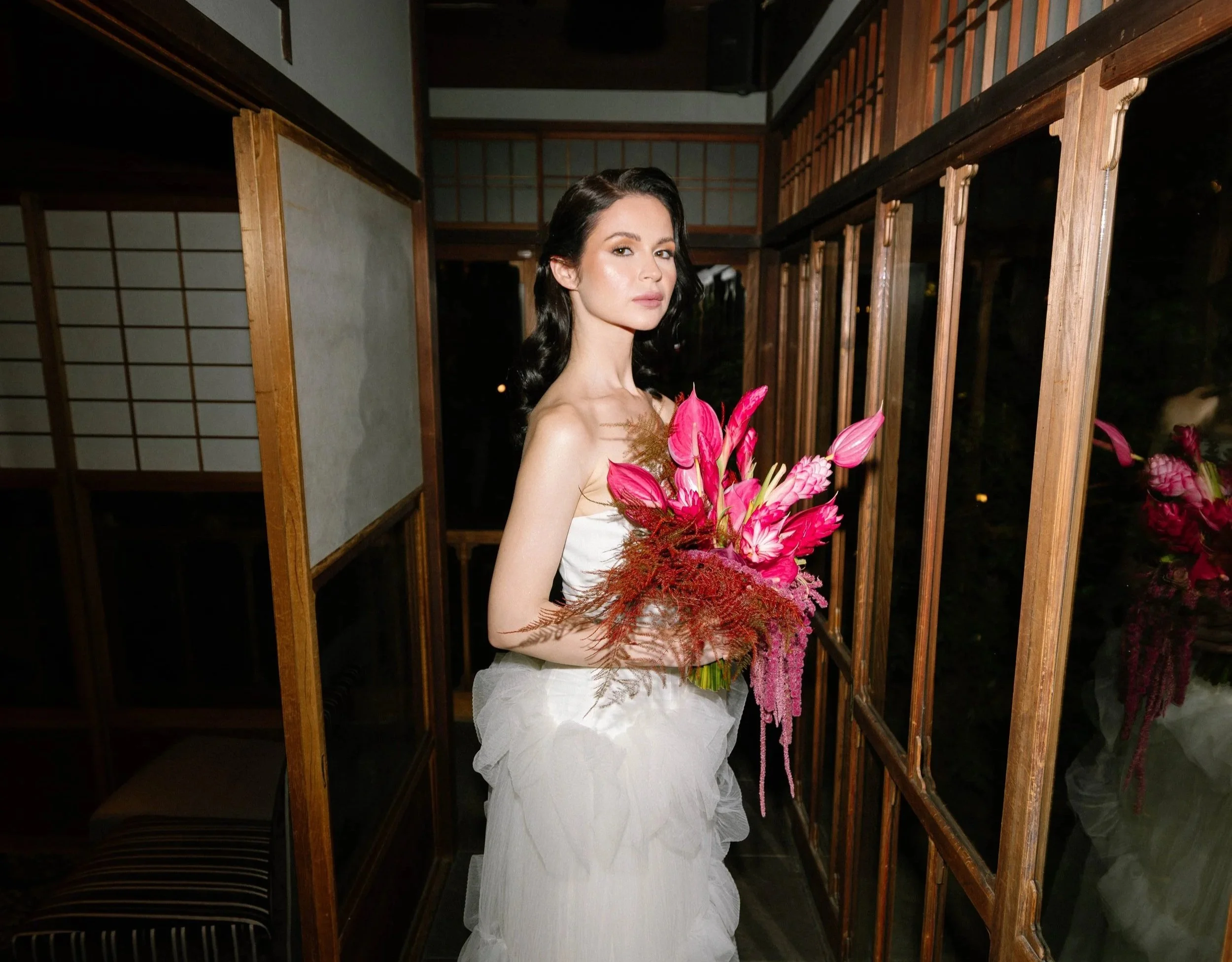 Bride alone in a dim traditional Japanese wooden corridor, holding a bouquet beneath a hanging lantern.