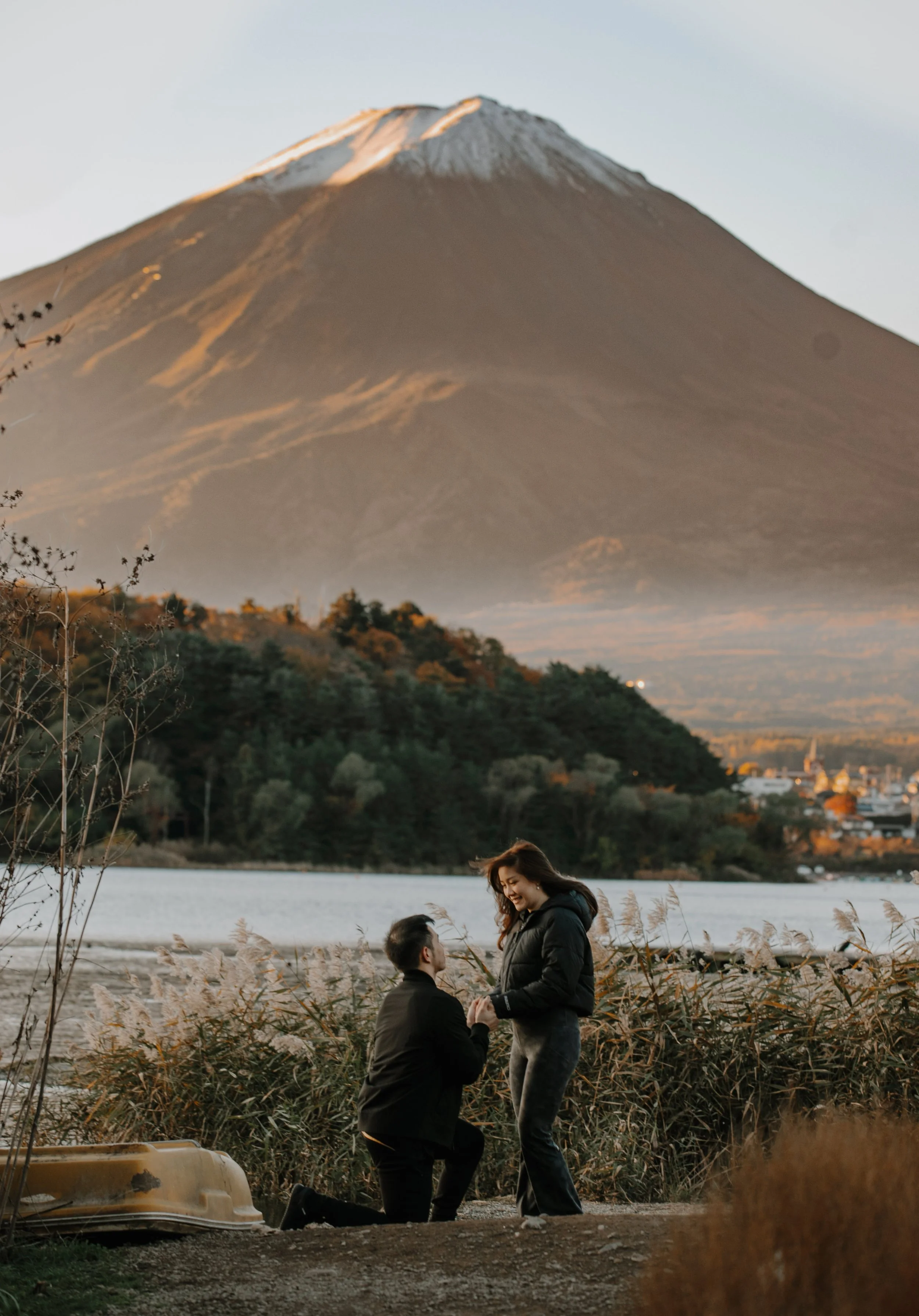 Man kneeling to propose to a laughing woman beside Lake Kawaguchiko at sunset with Mount Fuji behind