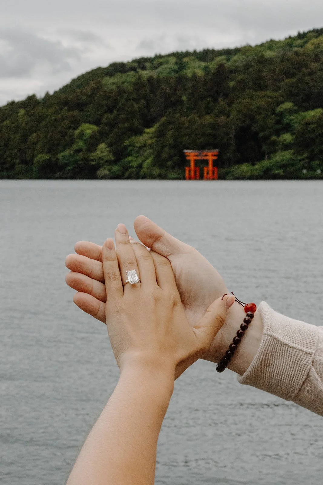 Close-up of hands revealing an emerald-cut engagement ring with the floating torii gate visible across Lake Ashi, Hakone