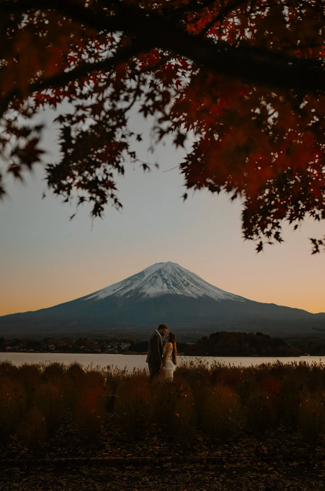 Couple kisses by the lake at sunset, framed beneath vivid red autumn maple leaves overhead.