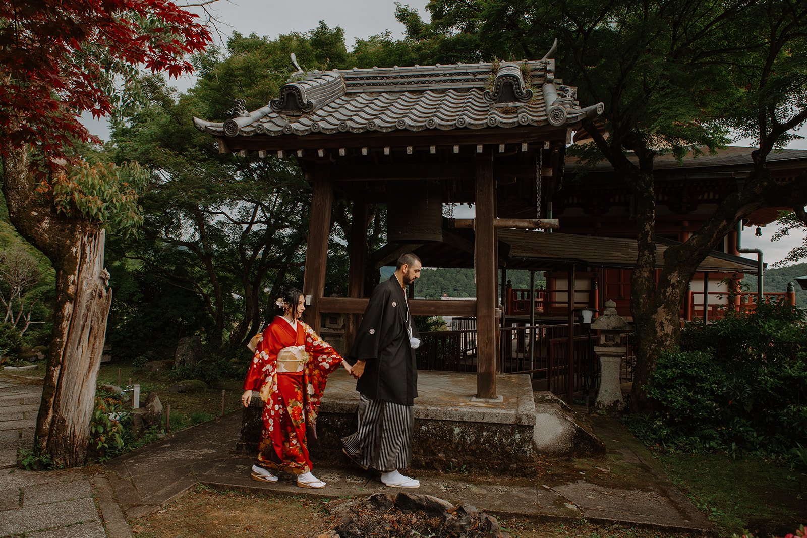 Couple in kimono walk hand in hand past a wooden temple bell tower overlooking the Kyoto hills.