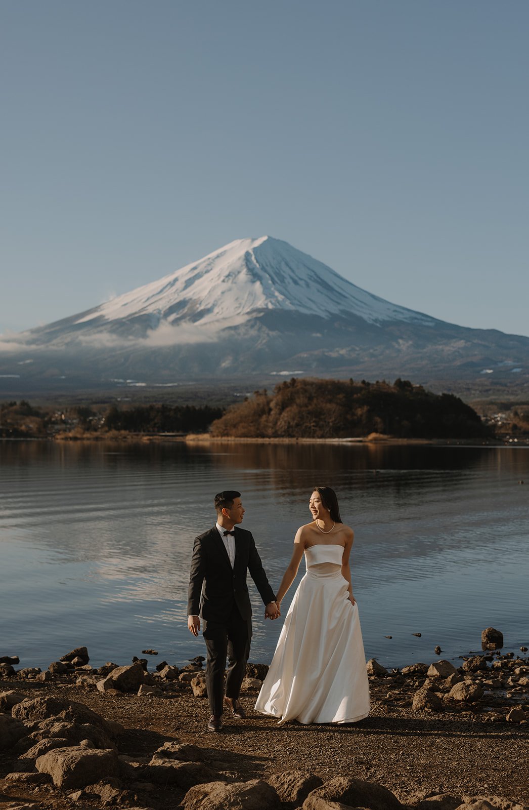Bride and groom walk together along the shore, smiling at each other beneath Mount Fuji.