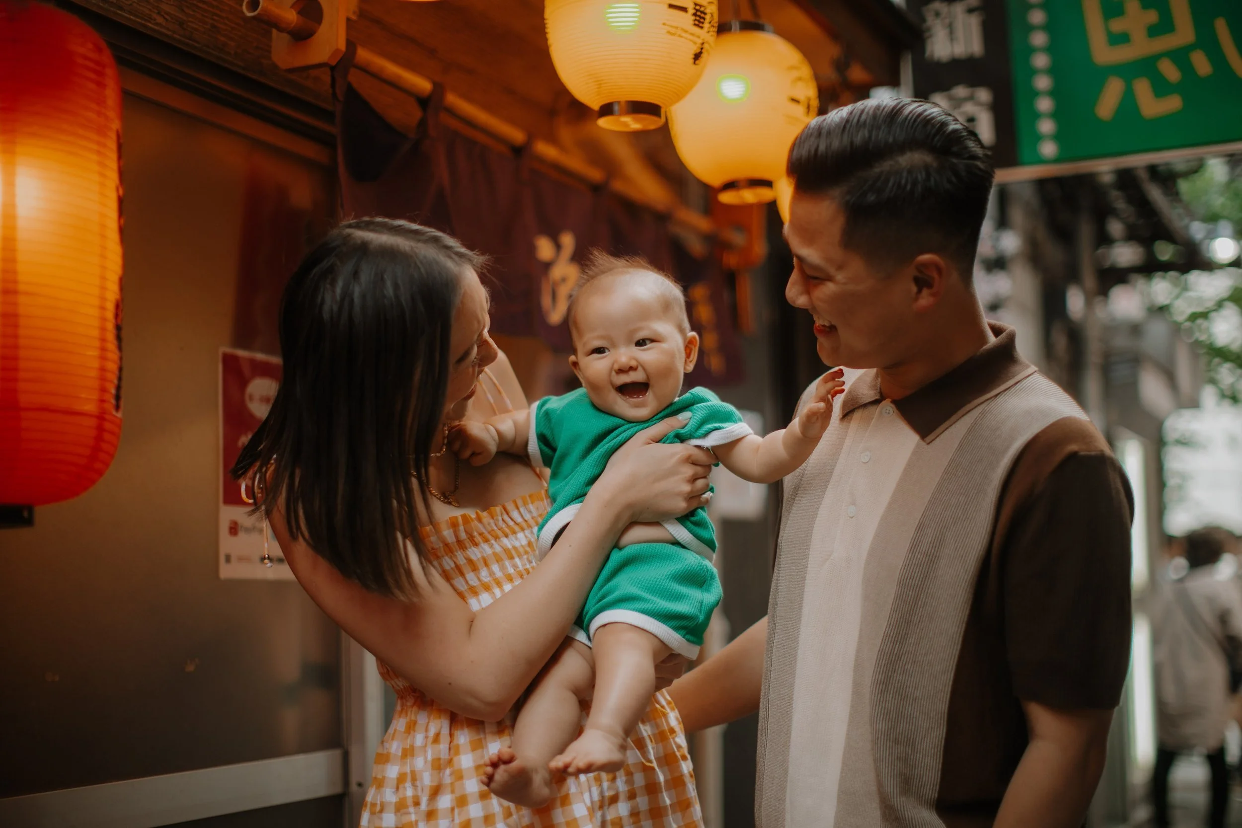 Parents laughing with their baby beneath a red paper lantern on a Tokyo backstreet during a family photography session.
