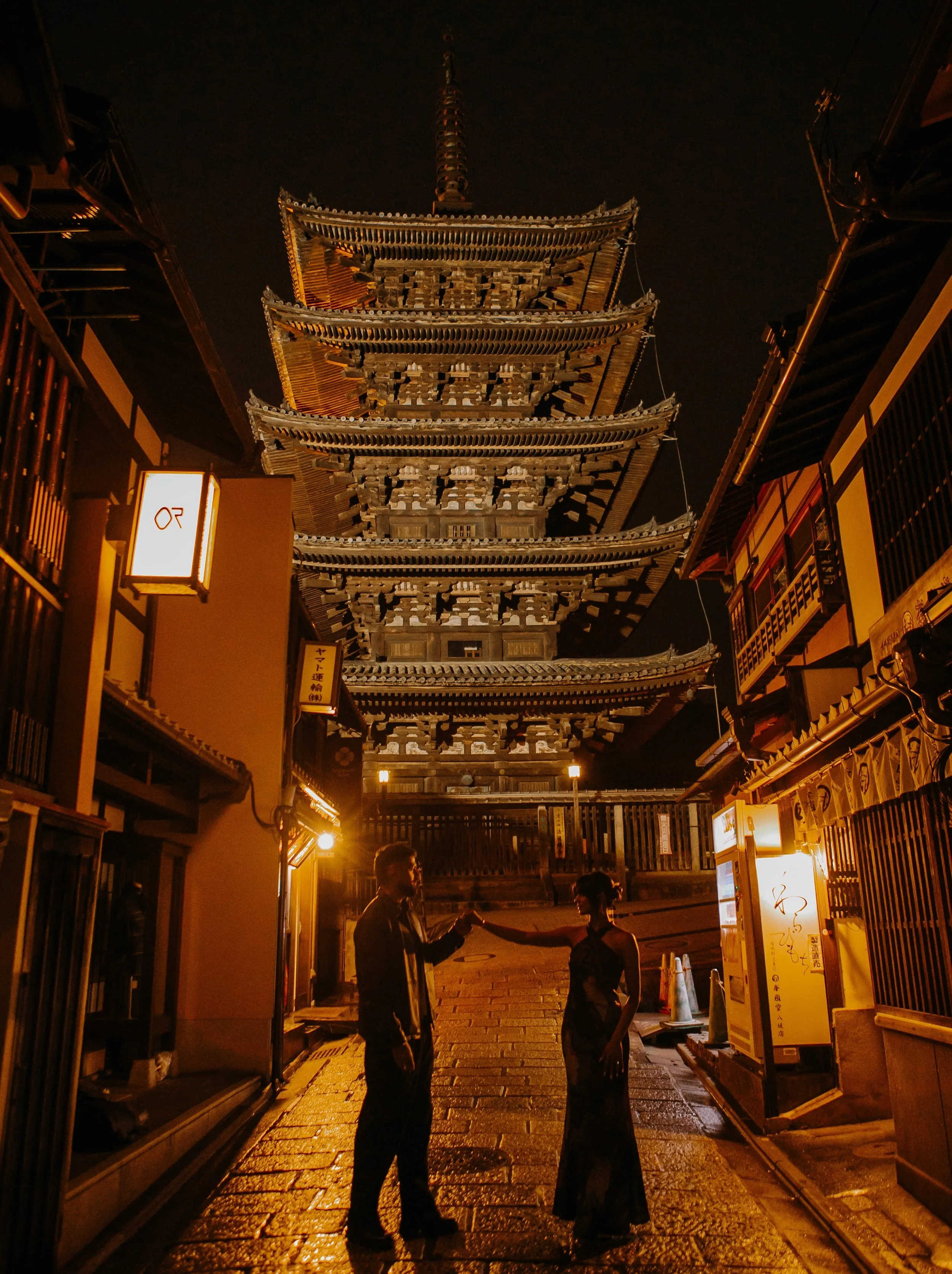 Couple dancing hand in hand on a rain-slicked lane beneath a five-story pagoda lit up at night.