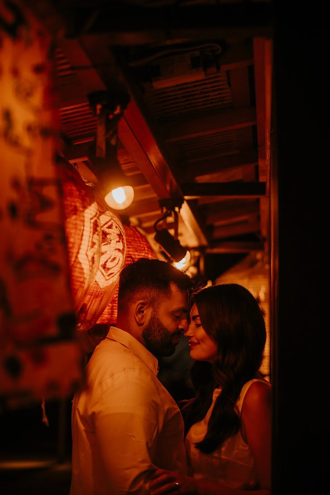 Engaged couple touching foreheads under a warm lantern in a narrow Tokyo izakaya alleyway