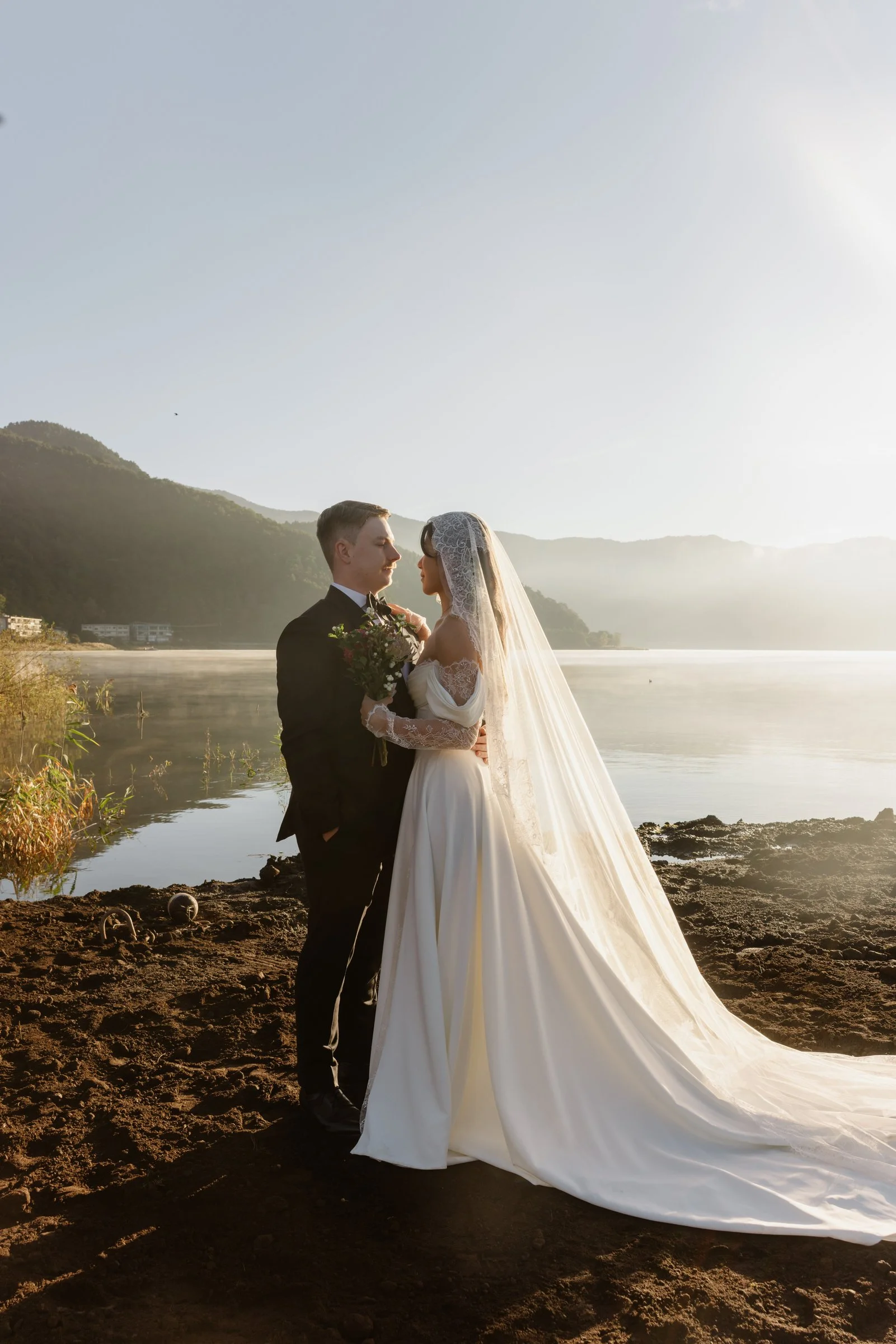 Bride in a long cathedral veil embracing her groom beside a misty lake at sunrise in Japan