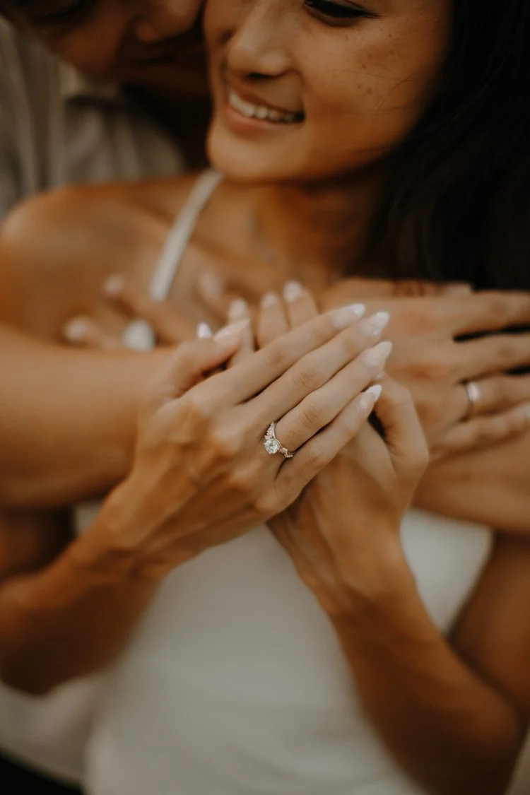 Close-up of a bride's hand with her engagement ring as her partner embraces her from behind