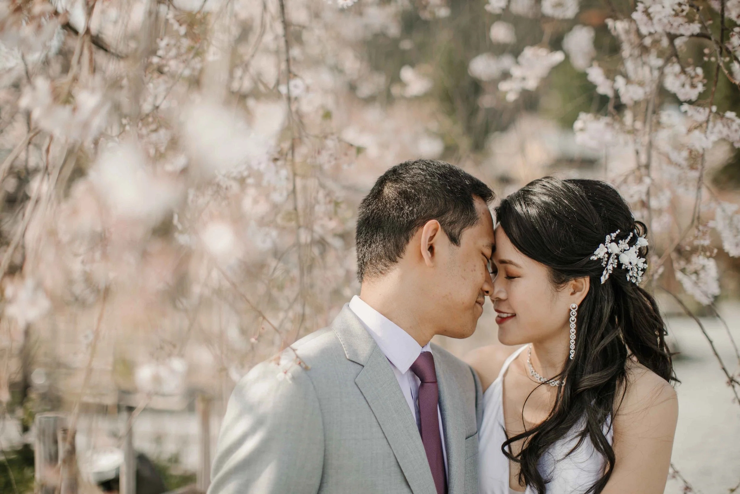 Couple embracing forehead to forehead beneath a weeping cherry blossom tree in Arashiyama, Kyoto, Japan, the bride wearing a floral hairpiece and diamond jewelry