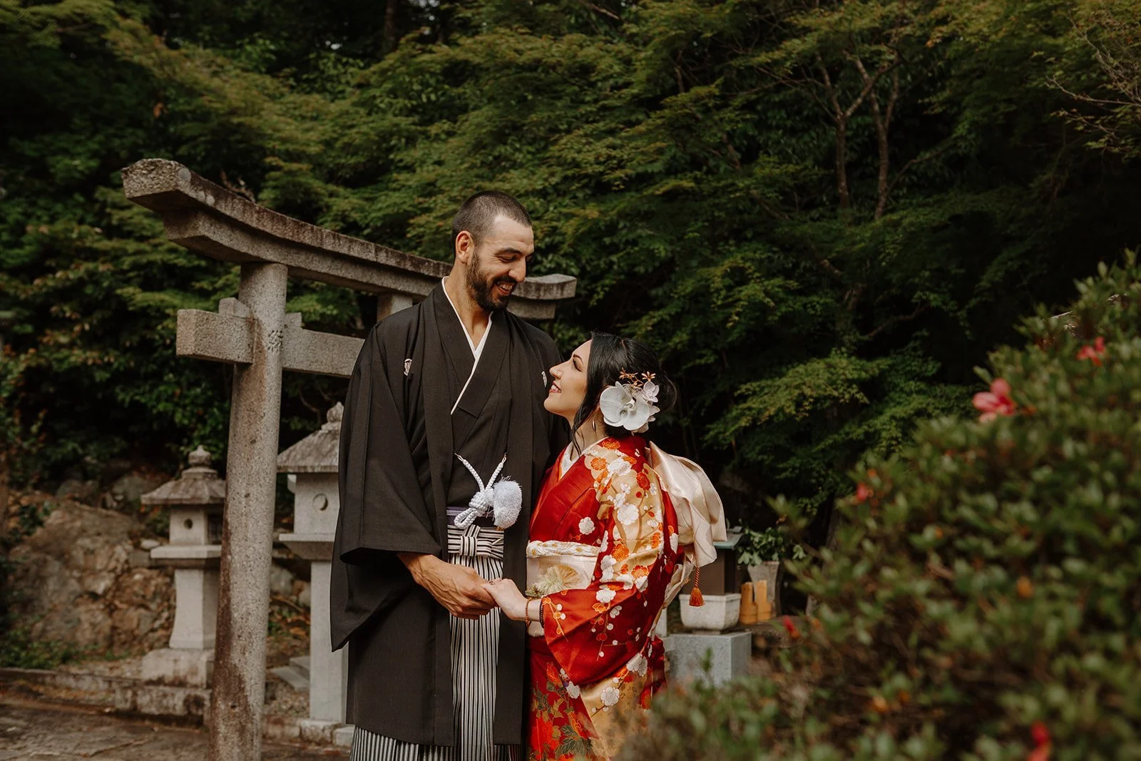 Couple in black and red kimono laughing together beside a stone torii gate surrounded by forest in Kyoto
