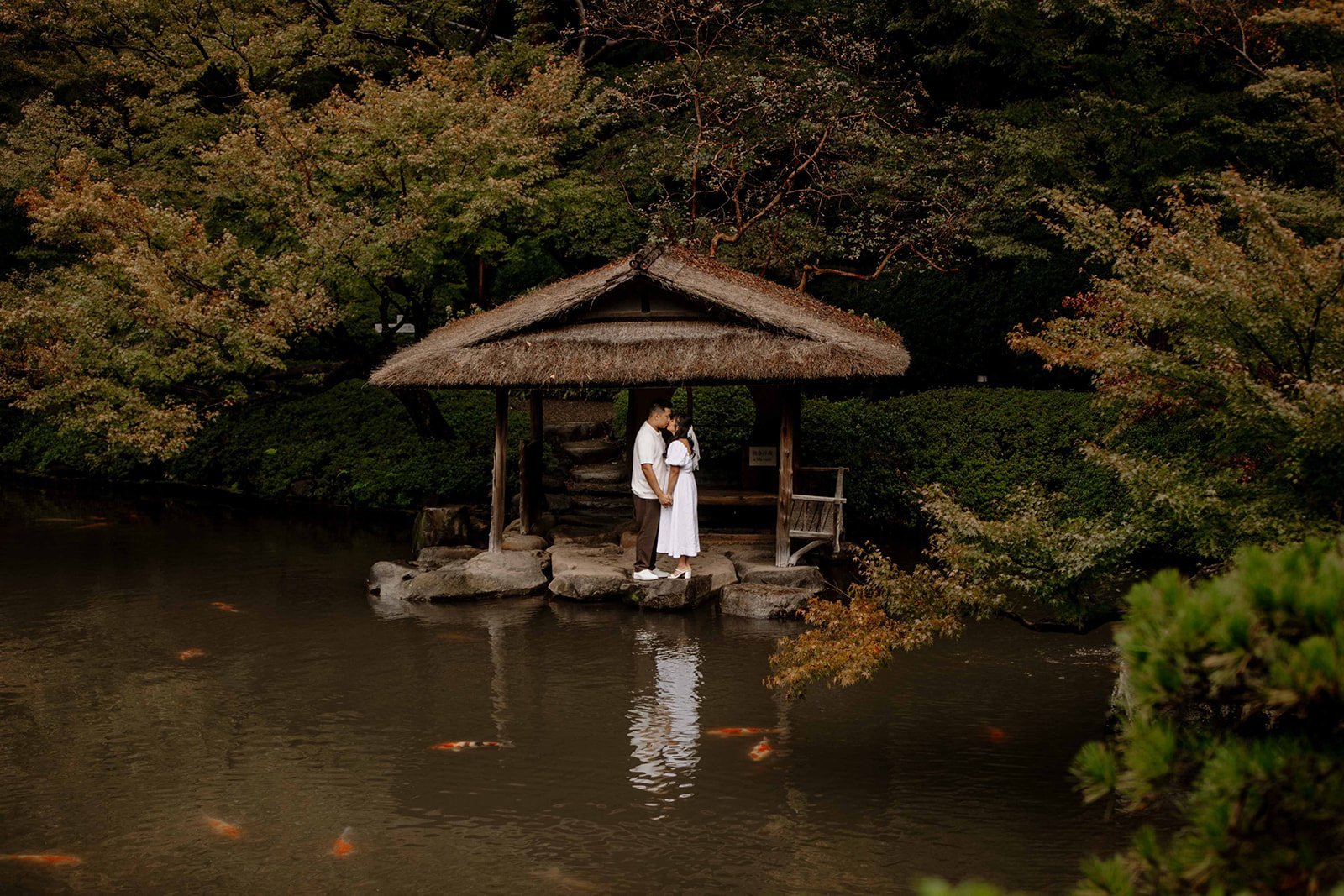Couple kissing under a thatched-roof teahouse pavilion beside a koi pond in a traditional Tokyo Japanese garden.